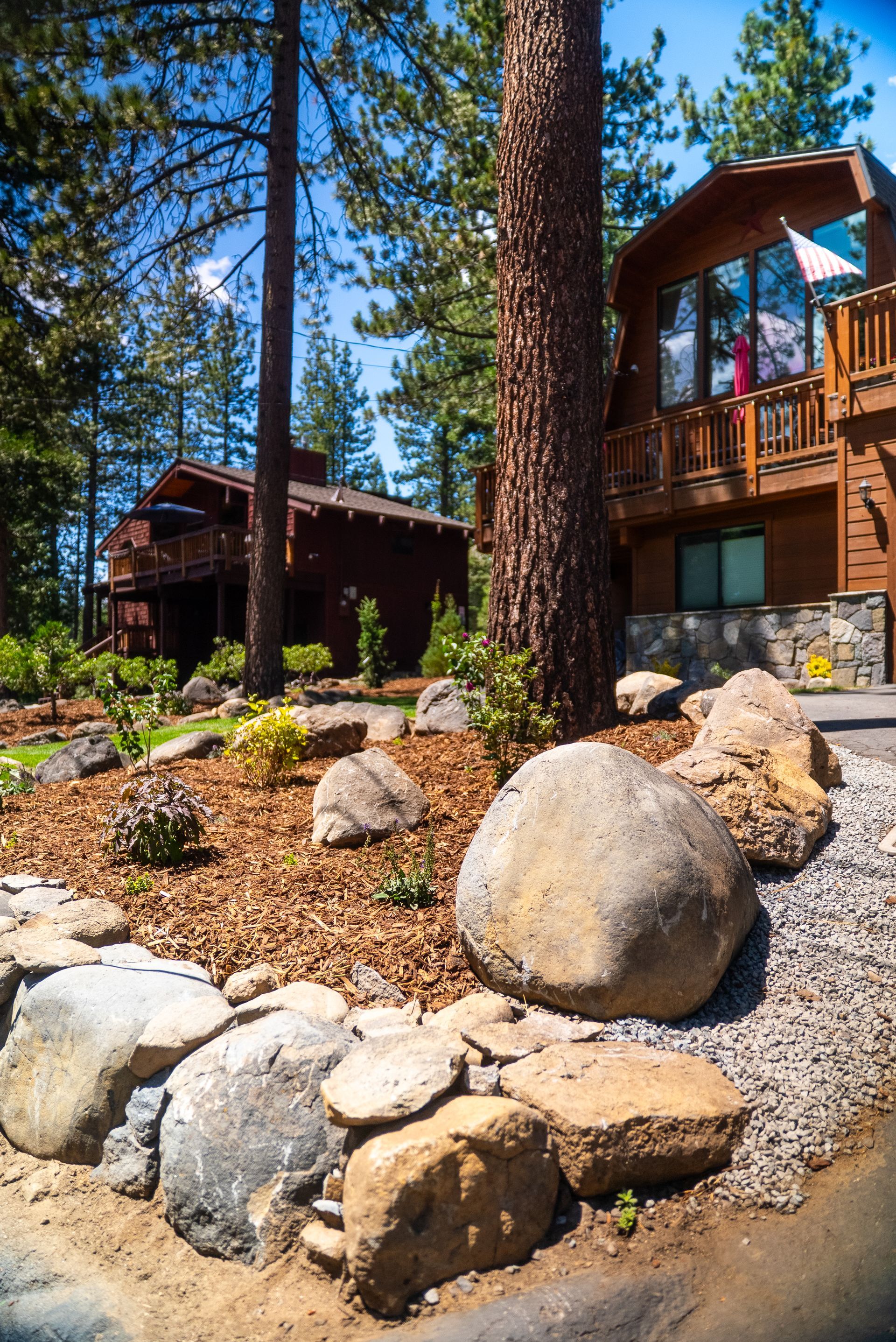 Stone-walled yard with large boulders and wood-sided buildings, a large tree, and landscaping.