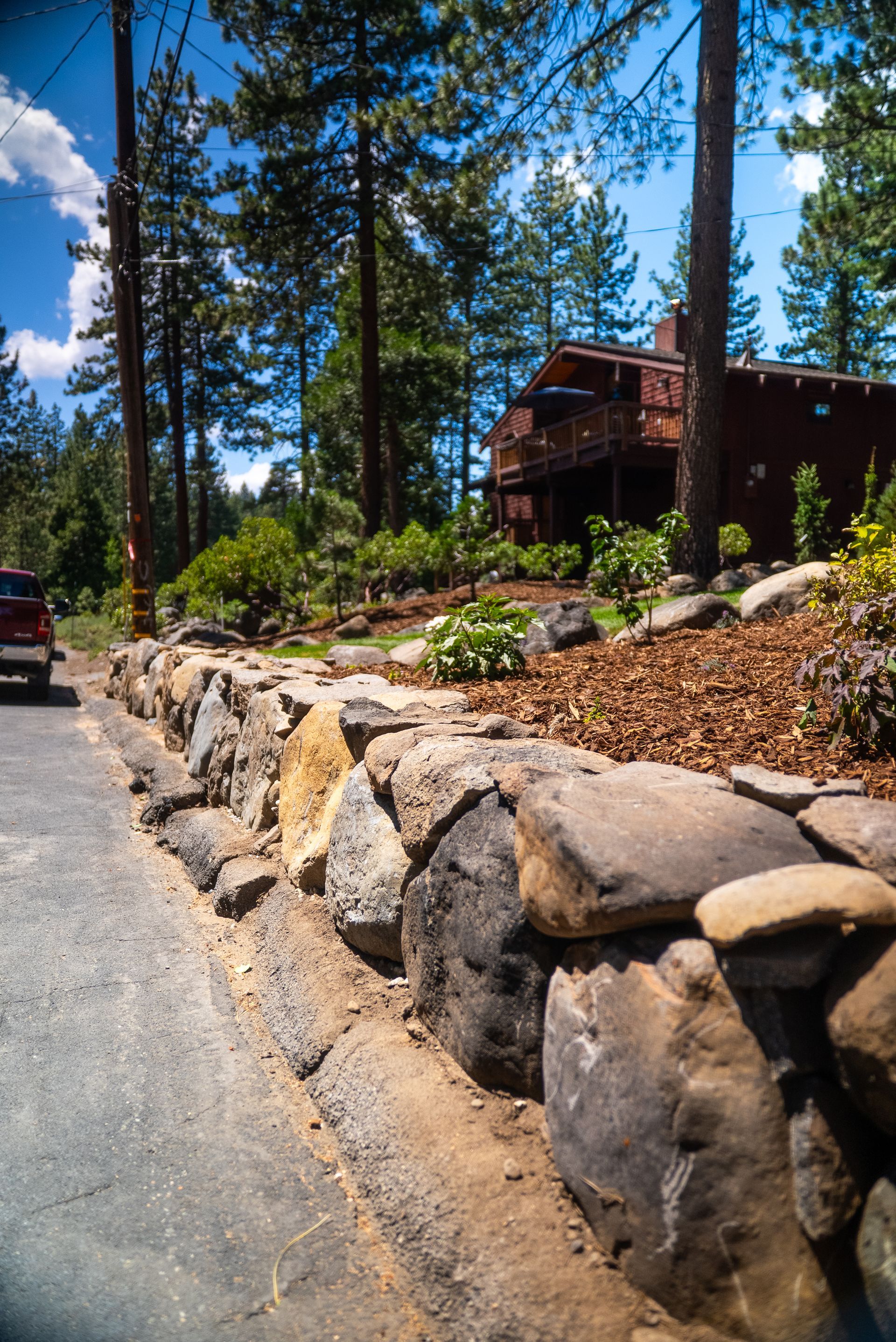 Stone retaining wall bordering a road, leading to a cabin in a forest.