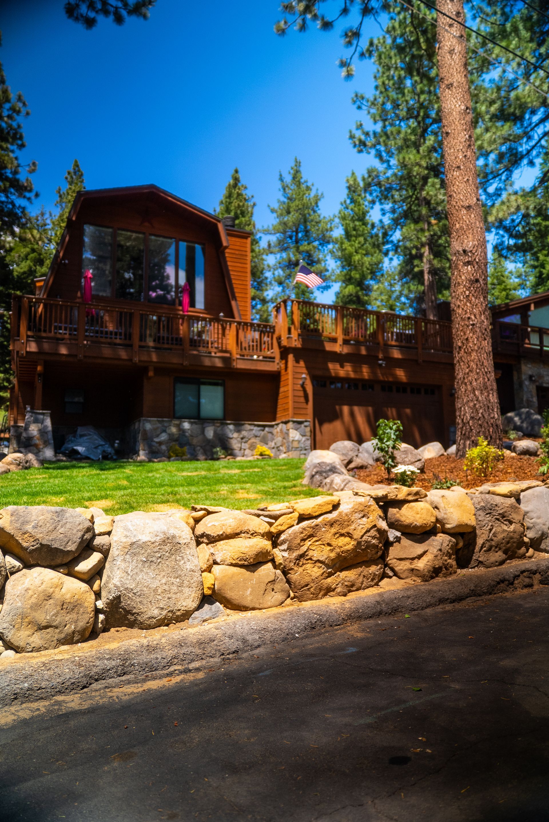 Wooden cabin with deck, stone wall, and green lawn under a sunny sky.
