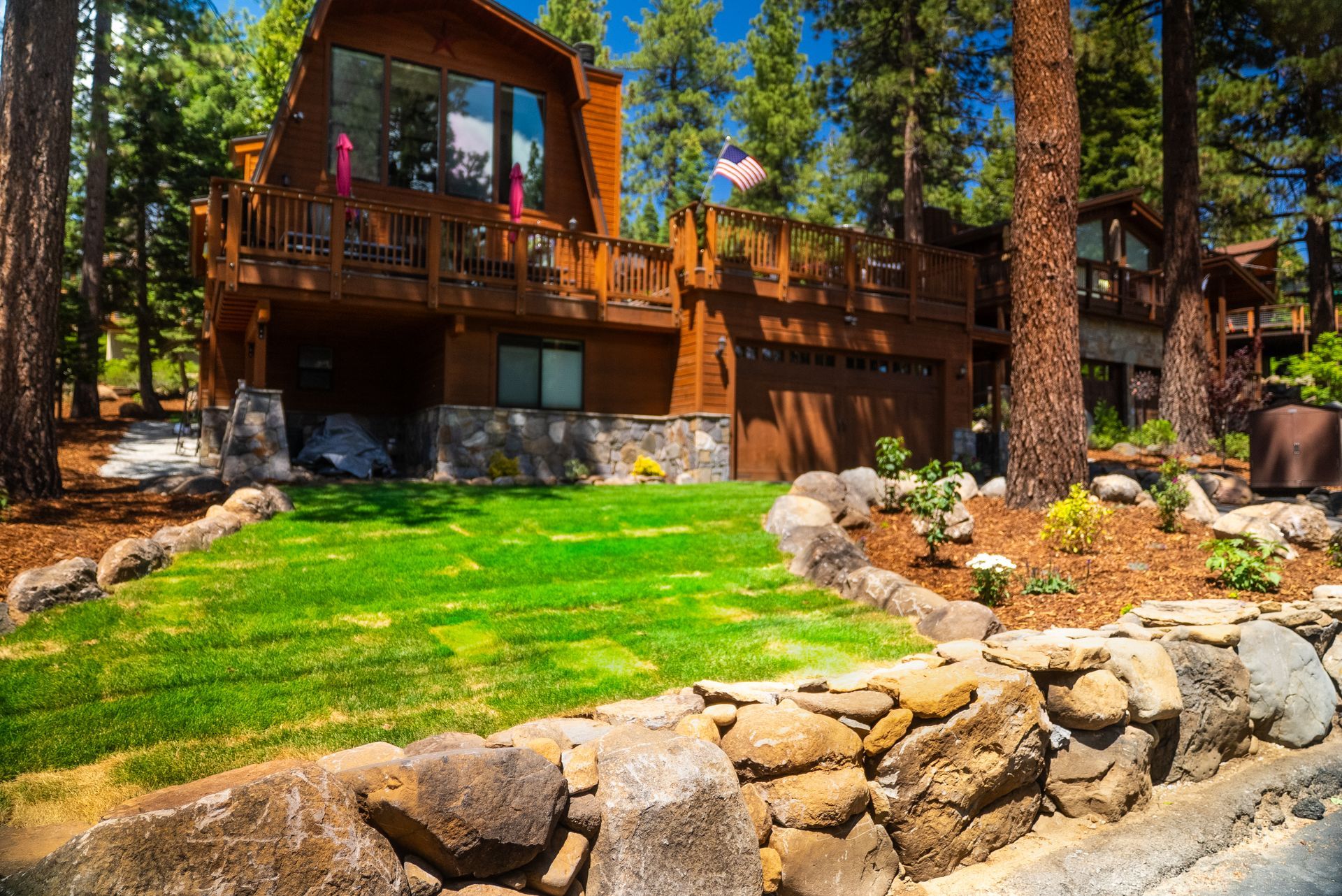 Brown wooden A-frame house with a green lawn, rock wall, and surrounding trees on a sunny day.