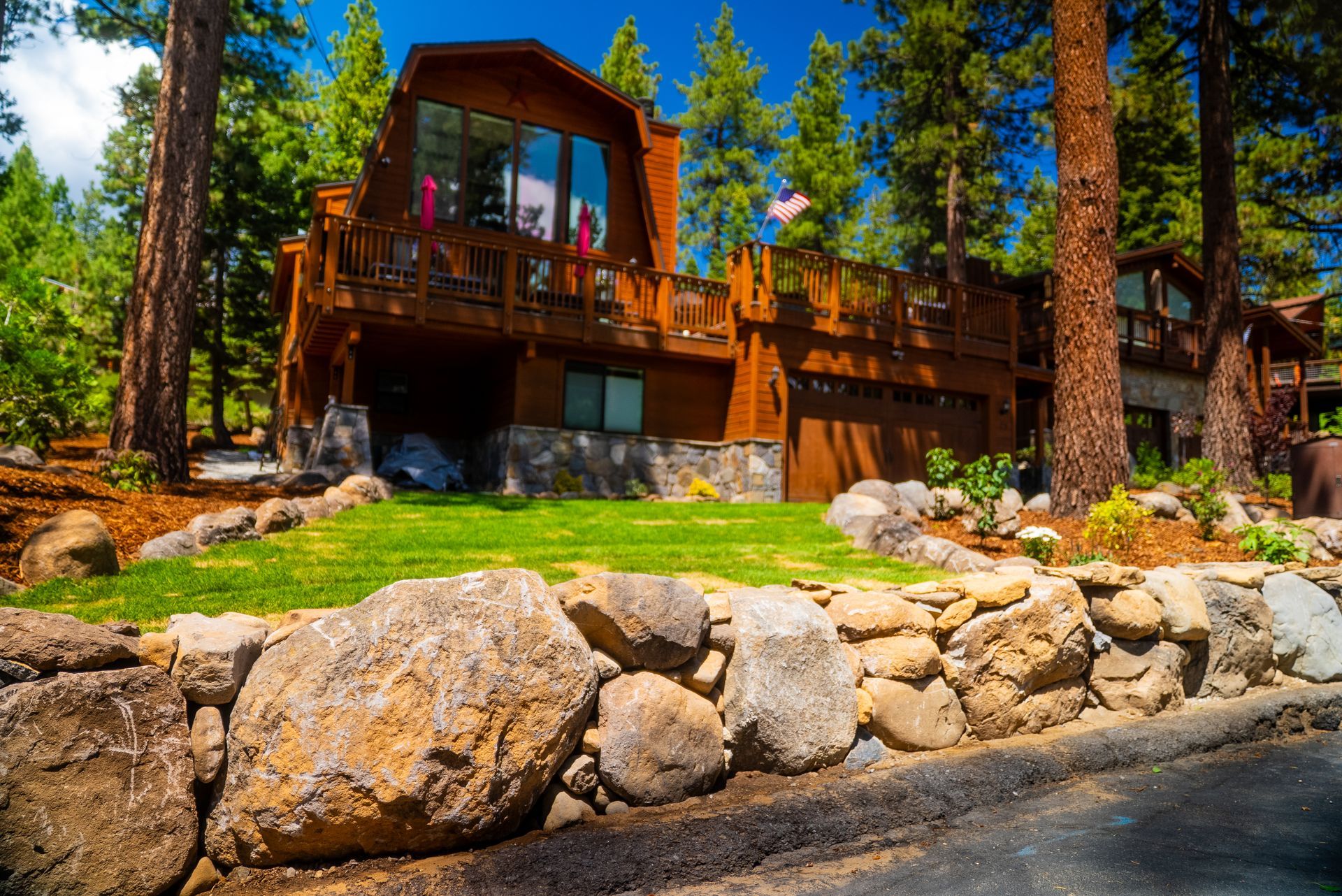 Brown wooden house with a balcony surrounded by tall trees and a rock wall in the foreground.