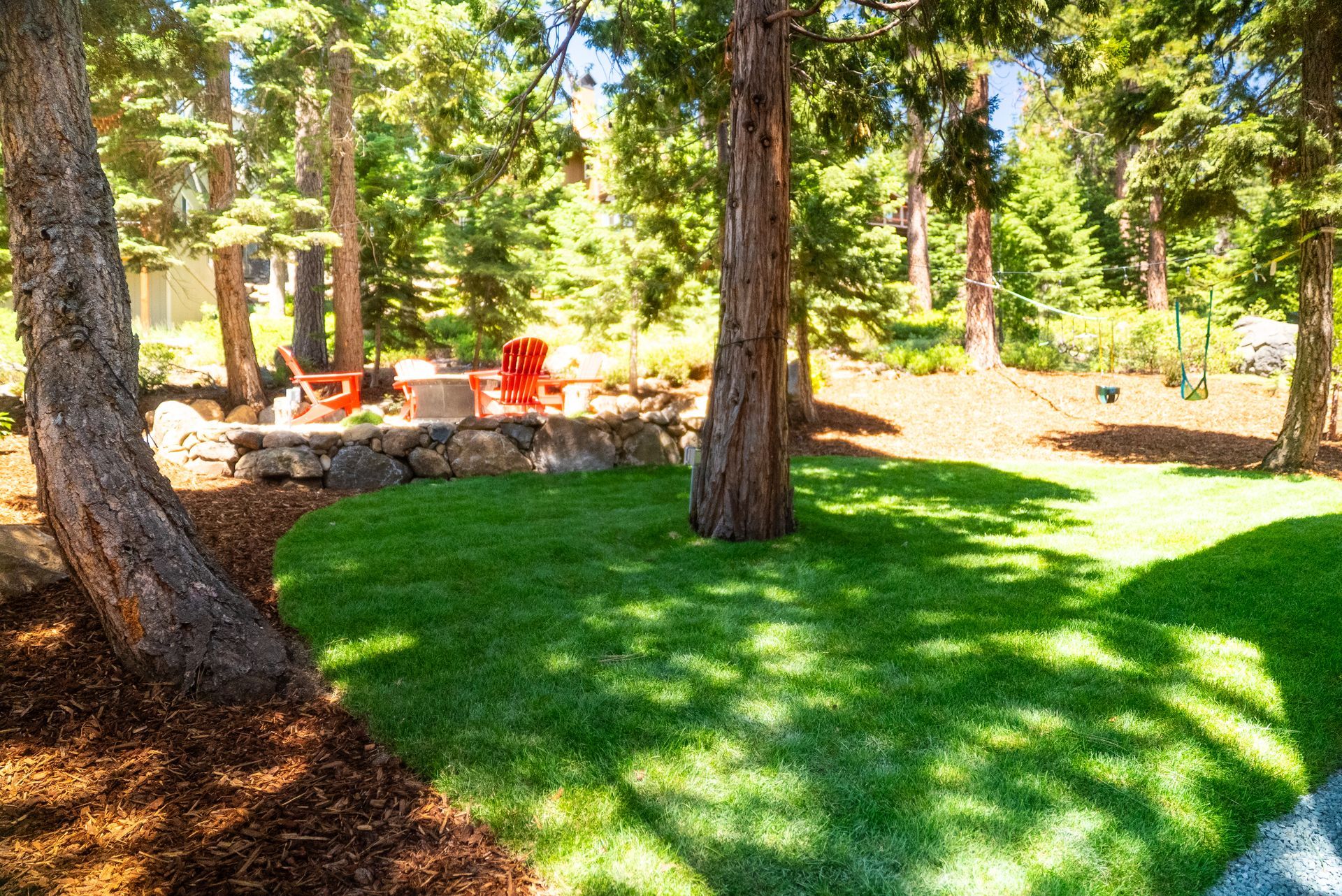 Lush green lawn with trees, a rock wall, and a fire pit area in a forest setting.