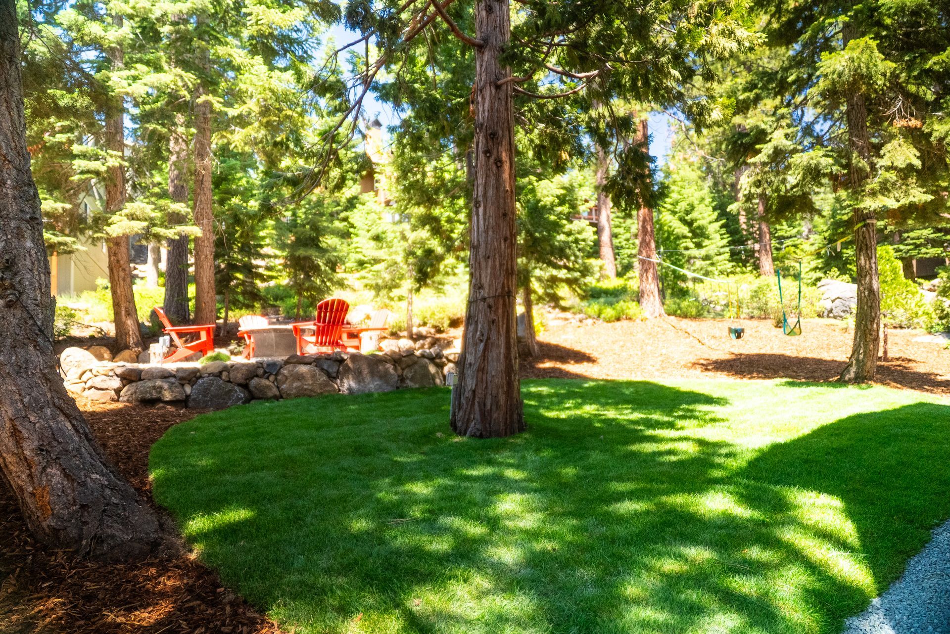 Lush green lawn with trees, stone wall, and red chairs in a sunny backyard.