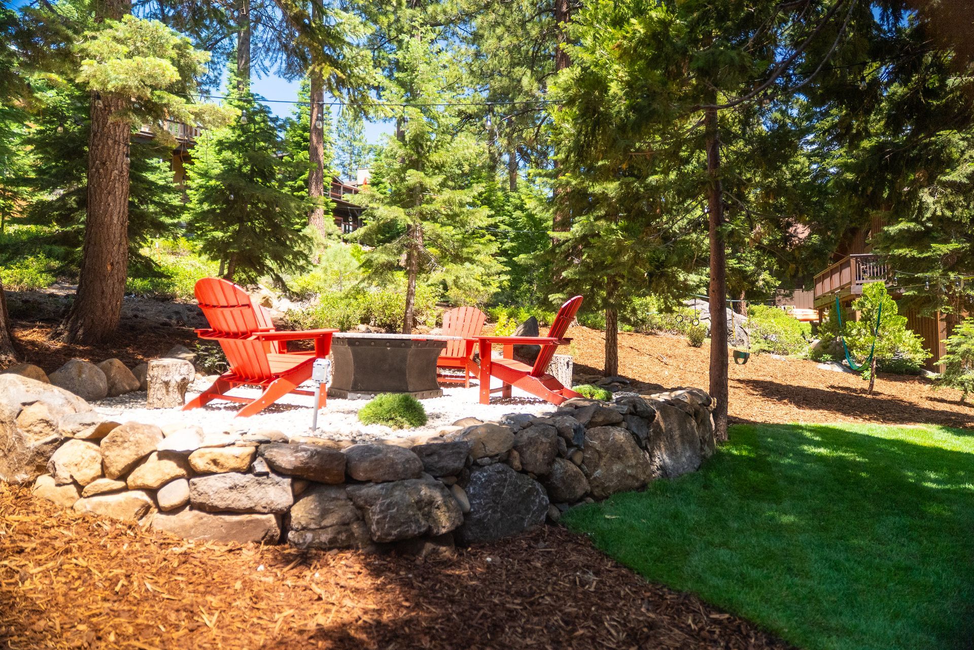 Outdoor seating area with fire pit, surrounded by a stone wall, lawn, and trees. Red Adirondack chairs.