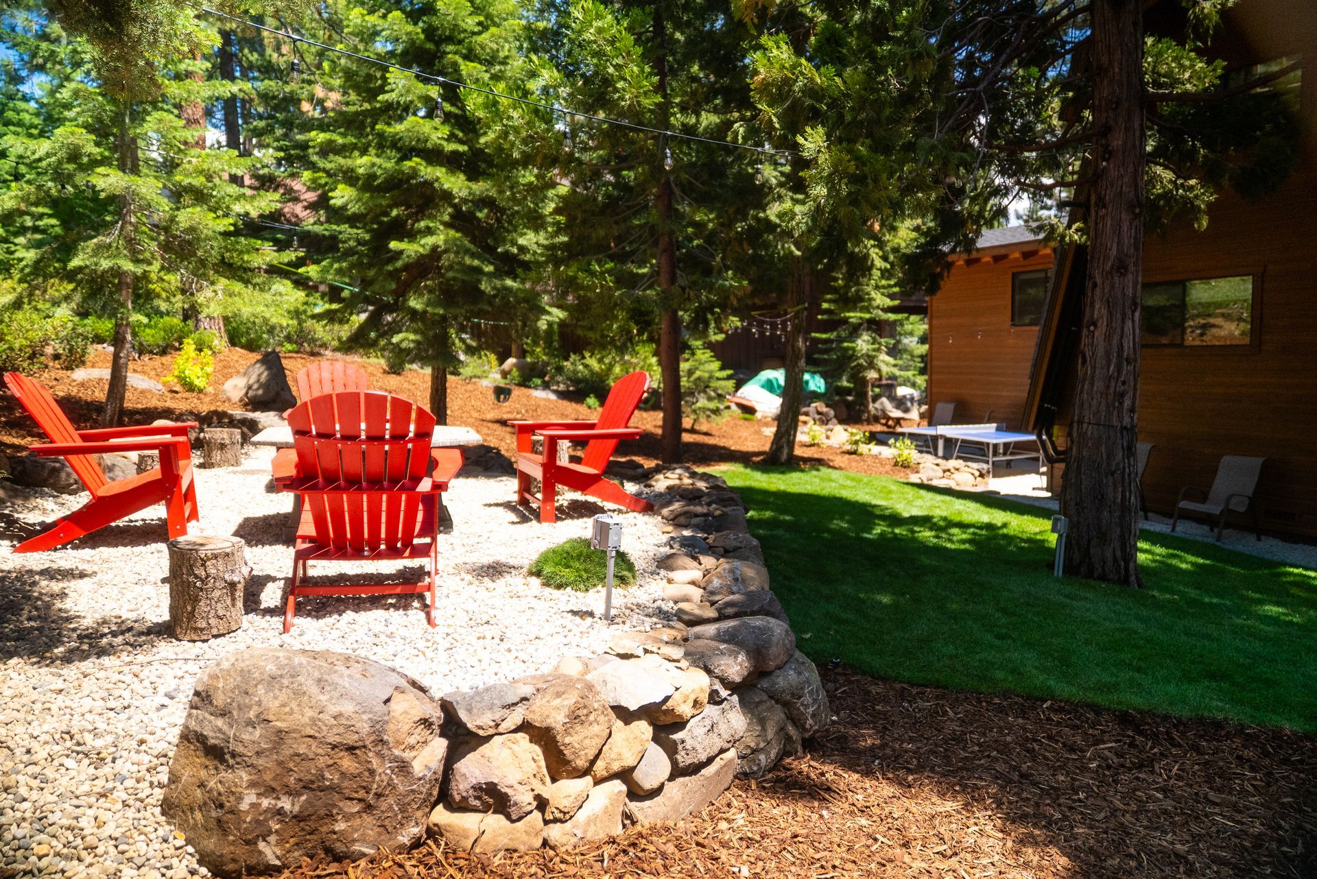 Red Adirondack chairs surround a small fire pit in a backyard with trees.