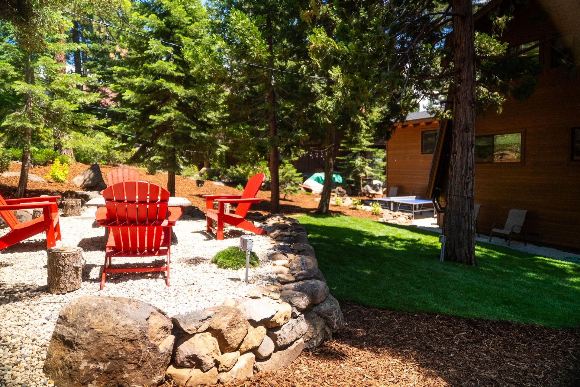 Red Adirondack chairs surround a fire pit on a gravel patio. Green lawn and trees surround the area.