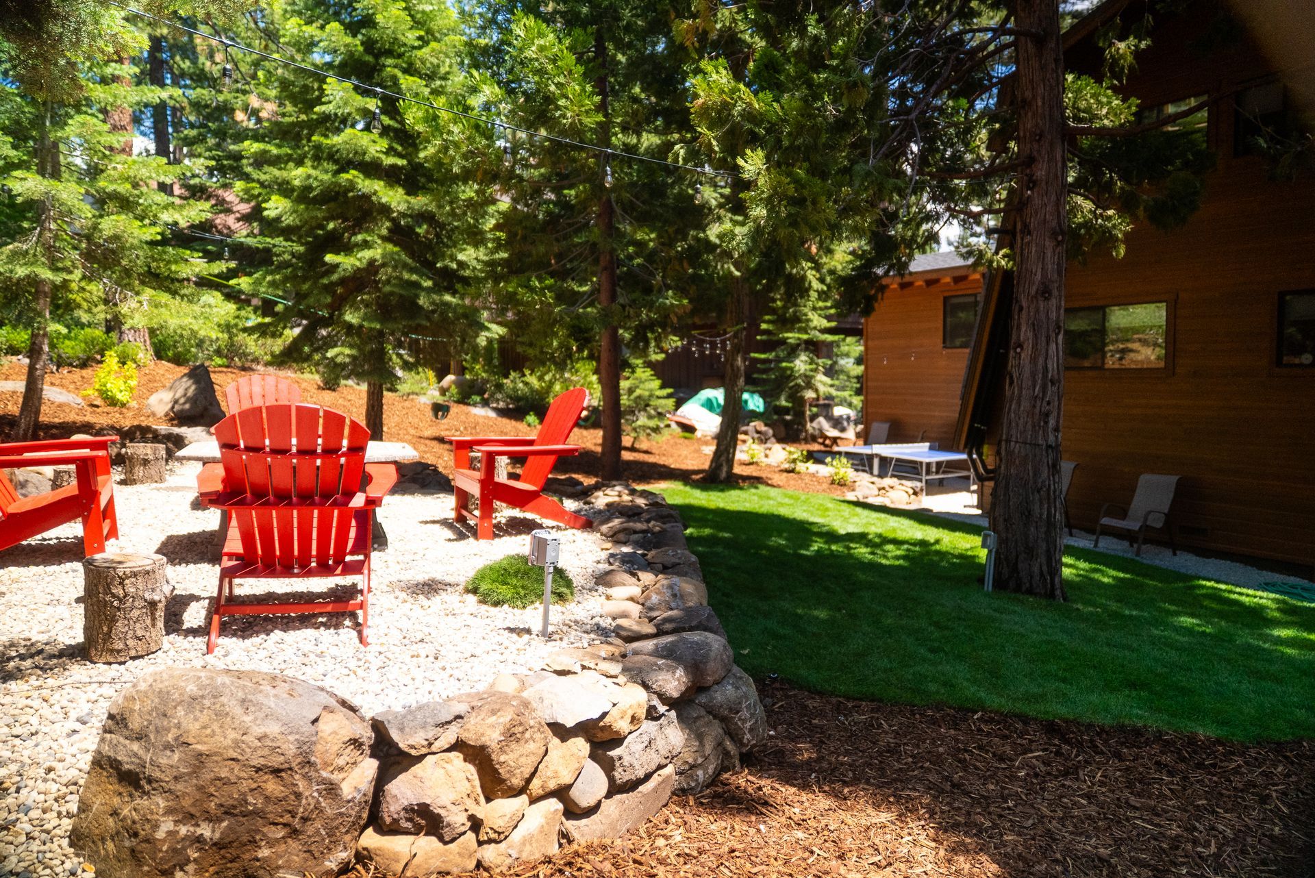Red Adirondack chairs surround a fire pit on a gravel patio, with a grassy lawn, trees, and a wooden building in the background.