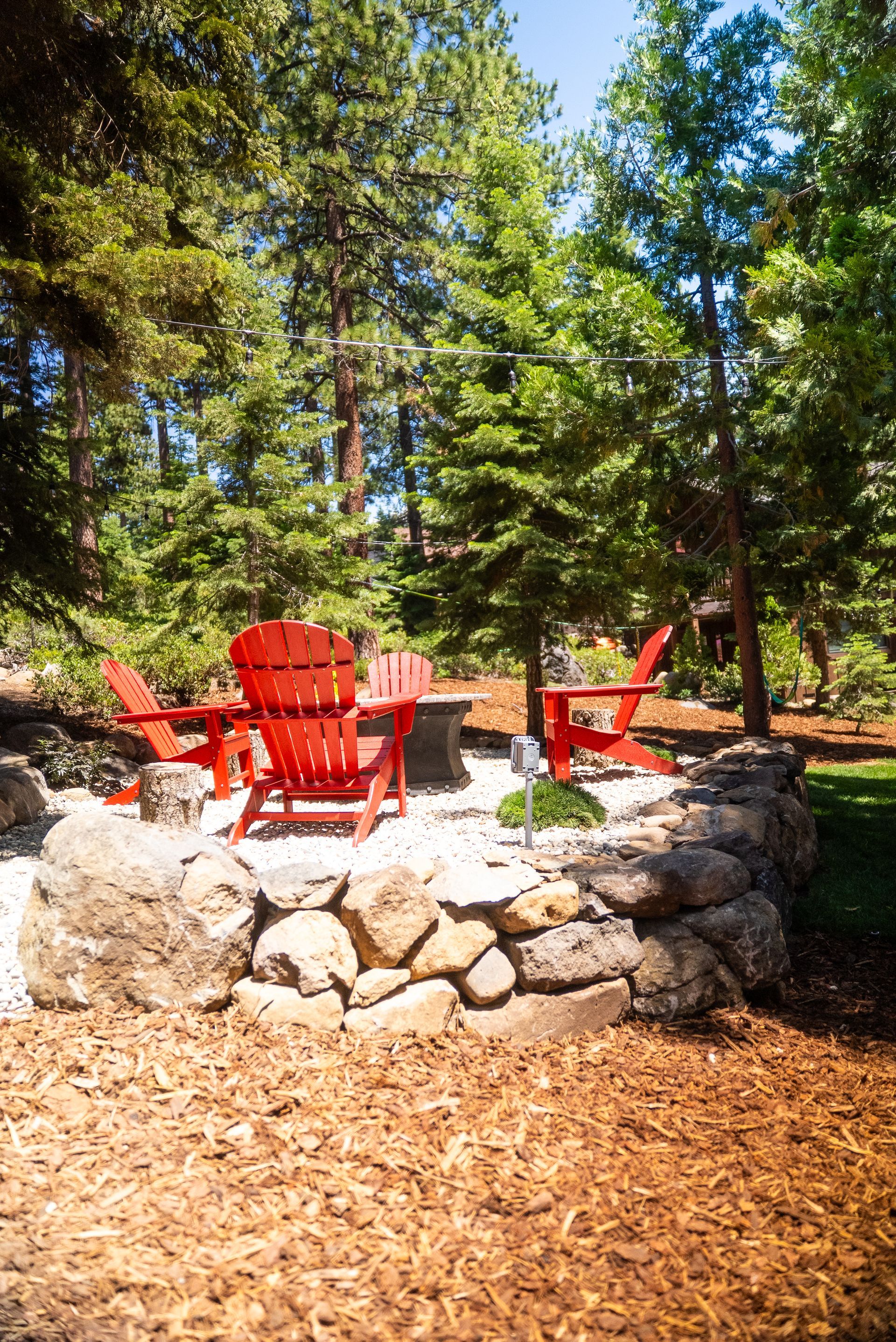 Red Adirondack chairs surround a fire pit on a stone patio, surrounded by trees.
