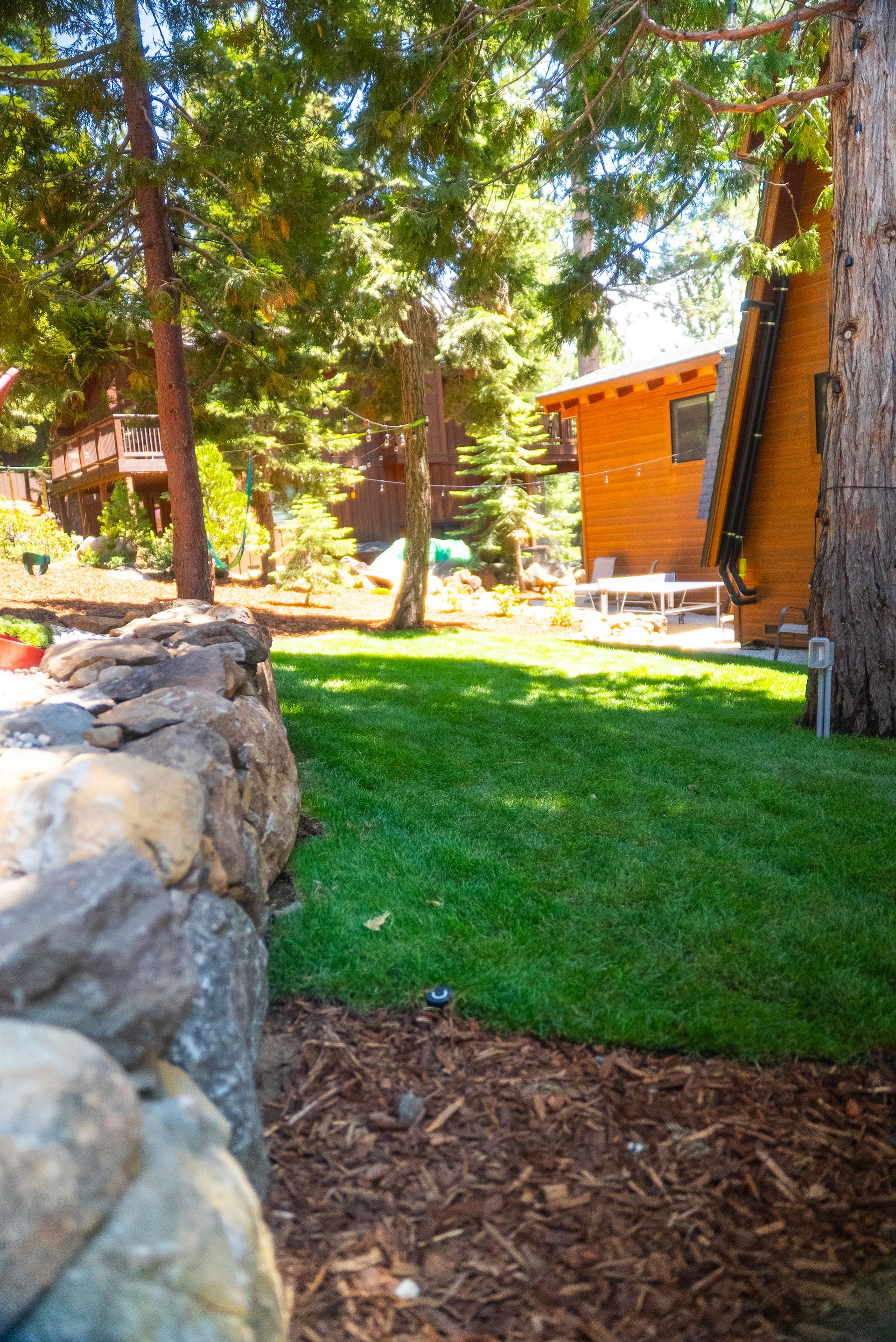 Lush green lawn with a stone wall in front of a wooden house, surrounded by trees.