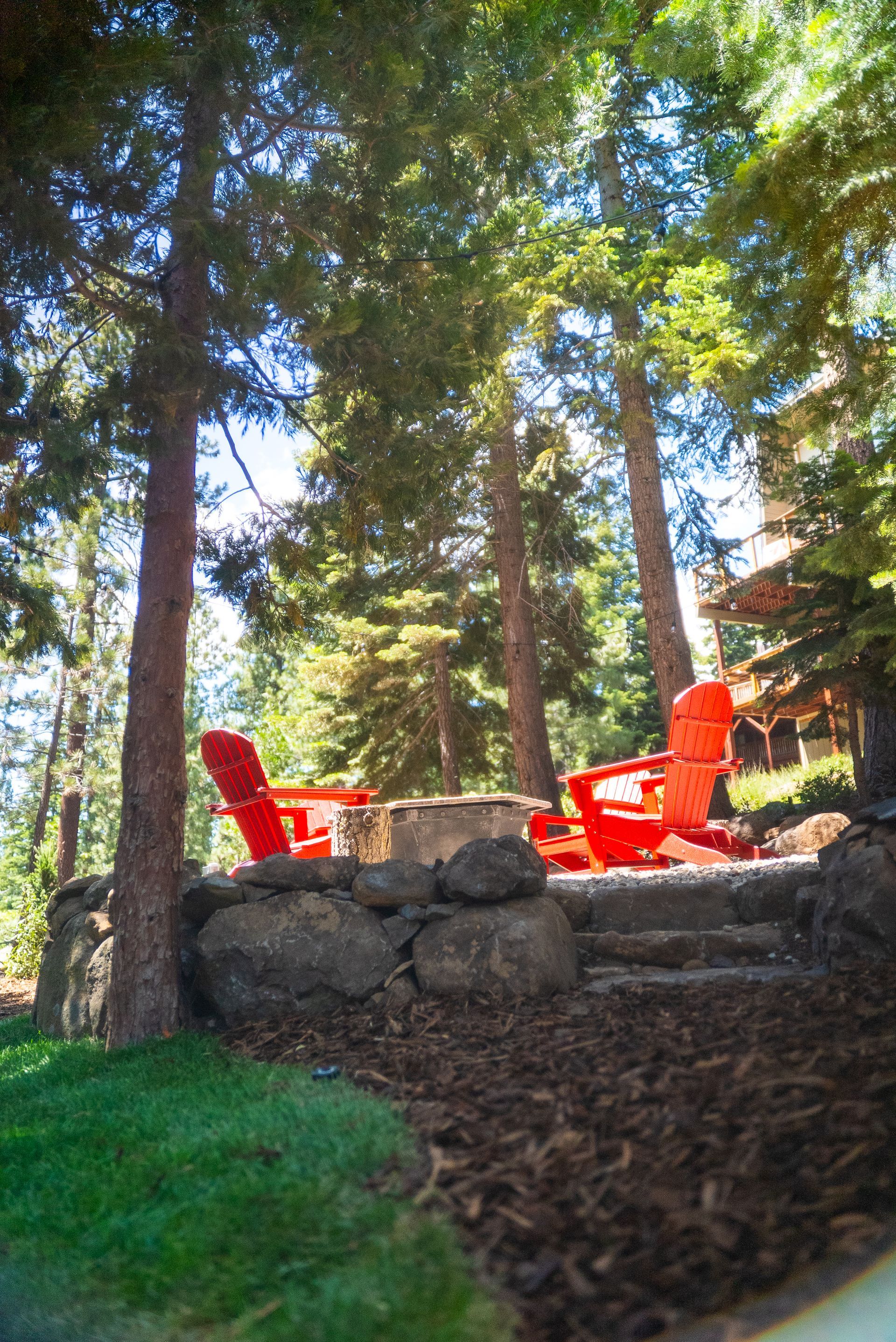 Red Adirondack chairs surround a stone fire pit in a wooded area.