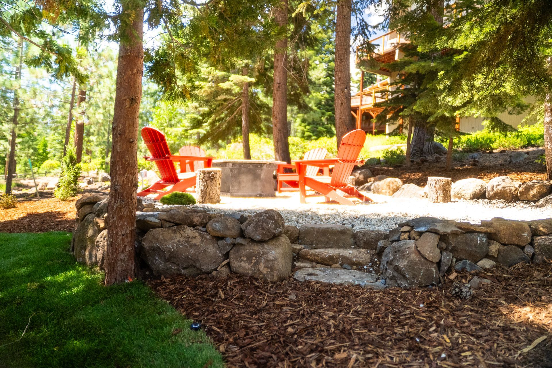 Fire pit with Adirondack chairs in a wooded setting; orange chairs, stone wall, and trees.