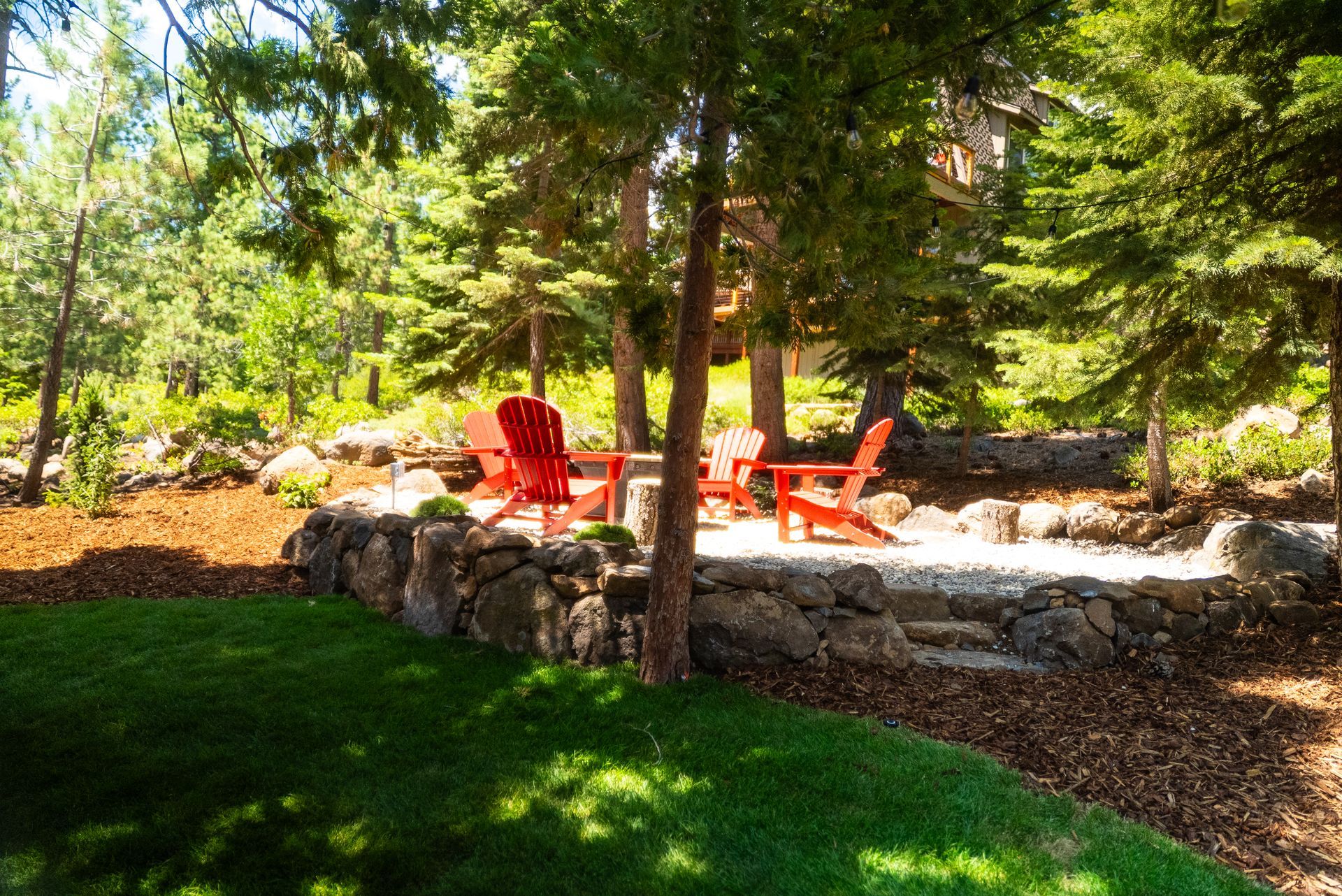Red Adirondack chairs surround a small table on a stone patio, surrounded by trees and greenery.