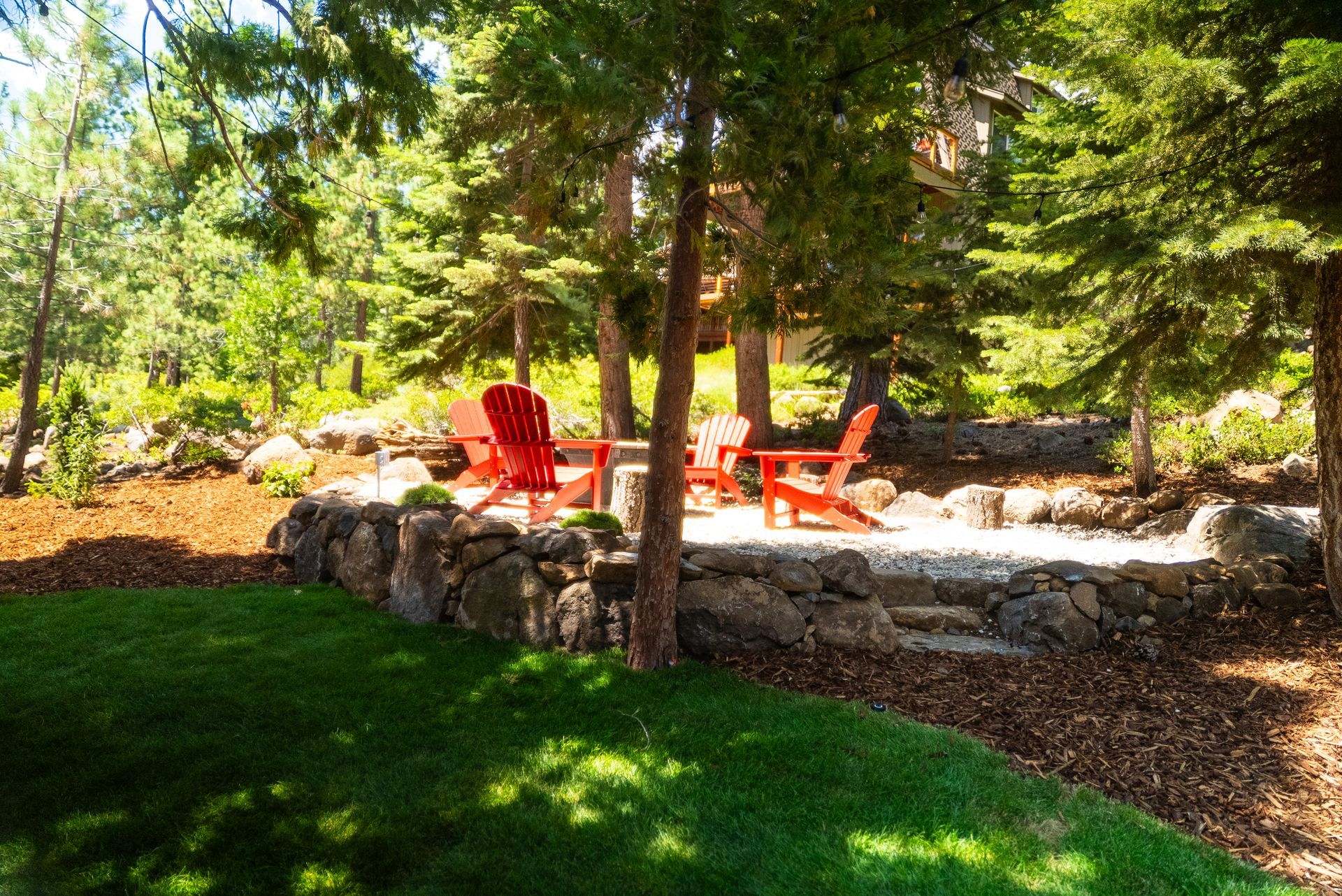 Lawn with a stone patio, orange Adirondack chairs under trees, surrounded by mulch and forest.