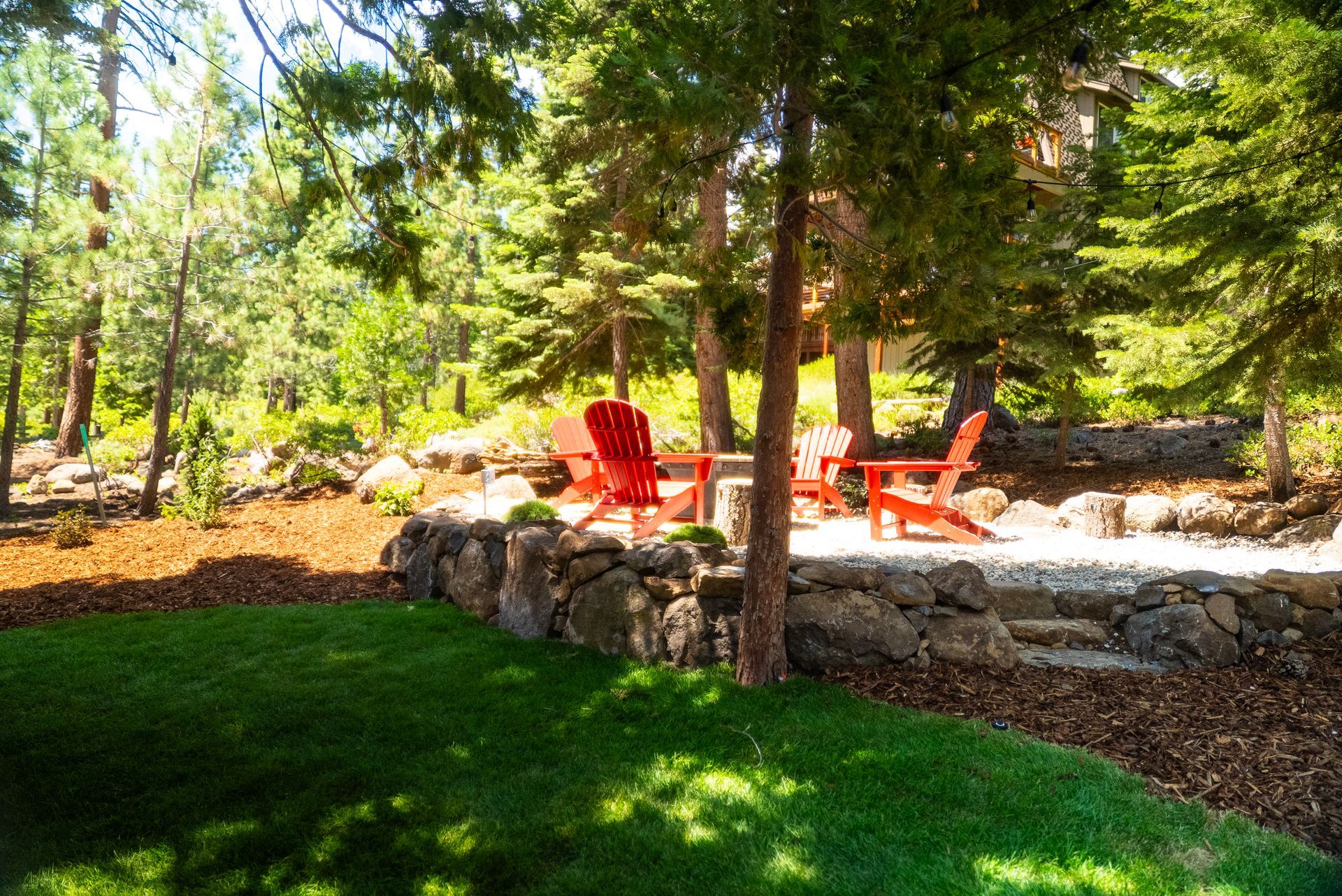 Red Adirondack chairs on stone patio surrounded by trees and a green lawn.