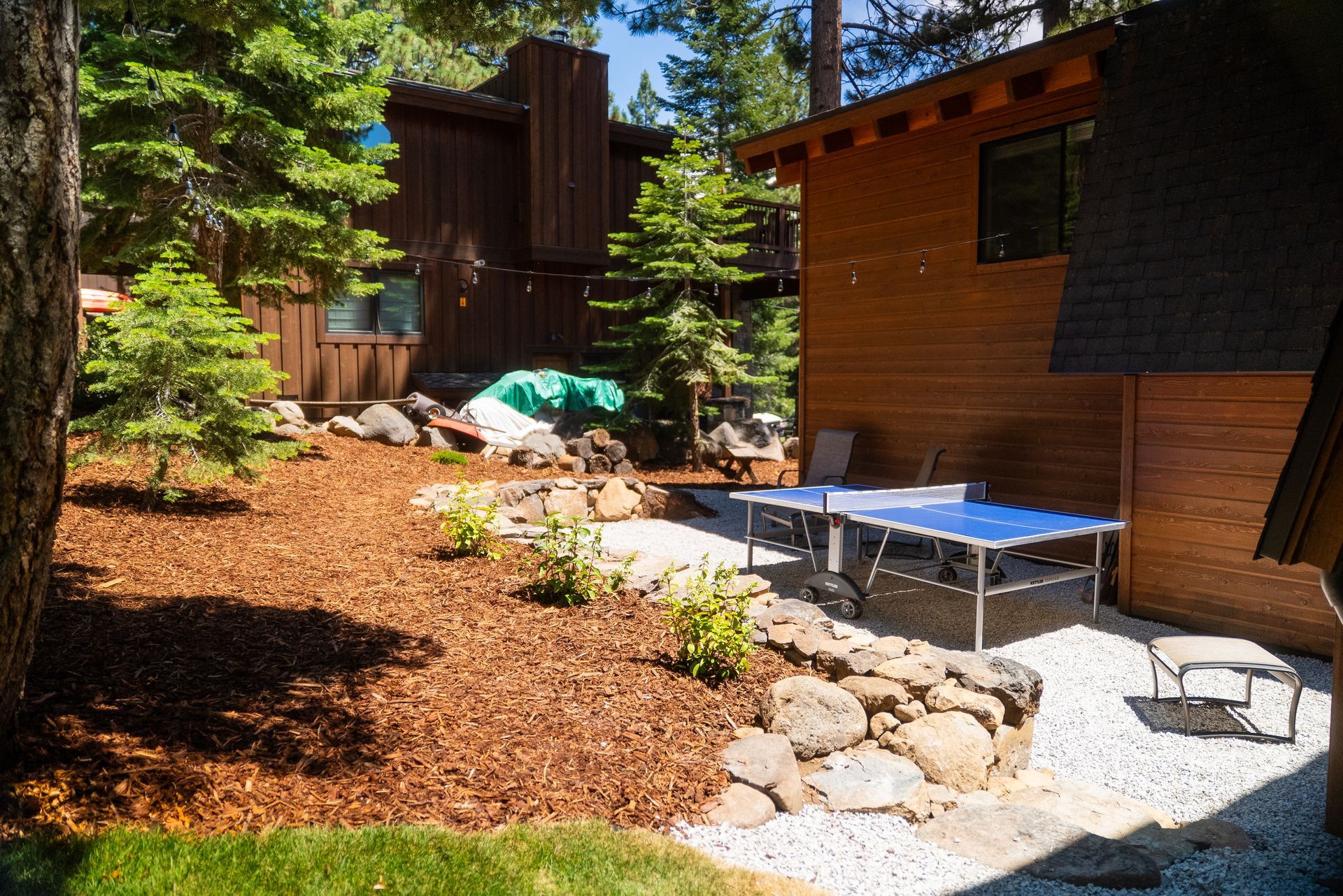 Backyard with a brown wood house and a ping pong table on a gravel patio area. Mulch and rocks are in the yard.