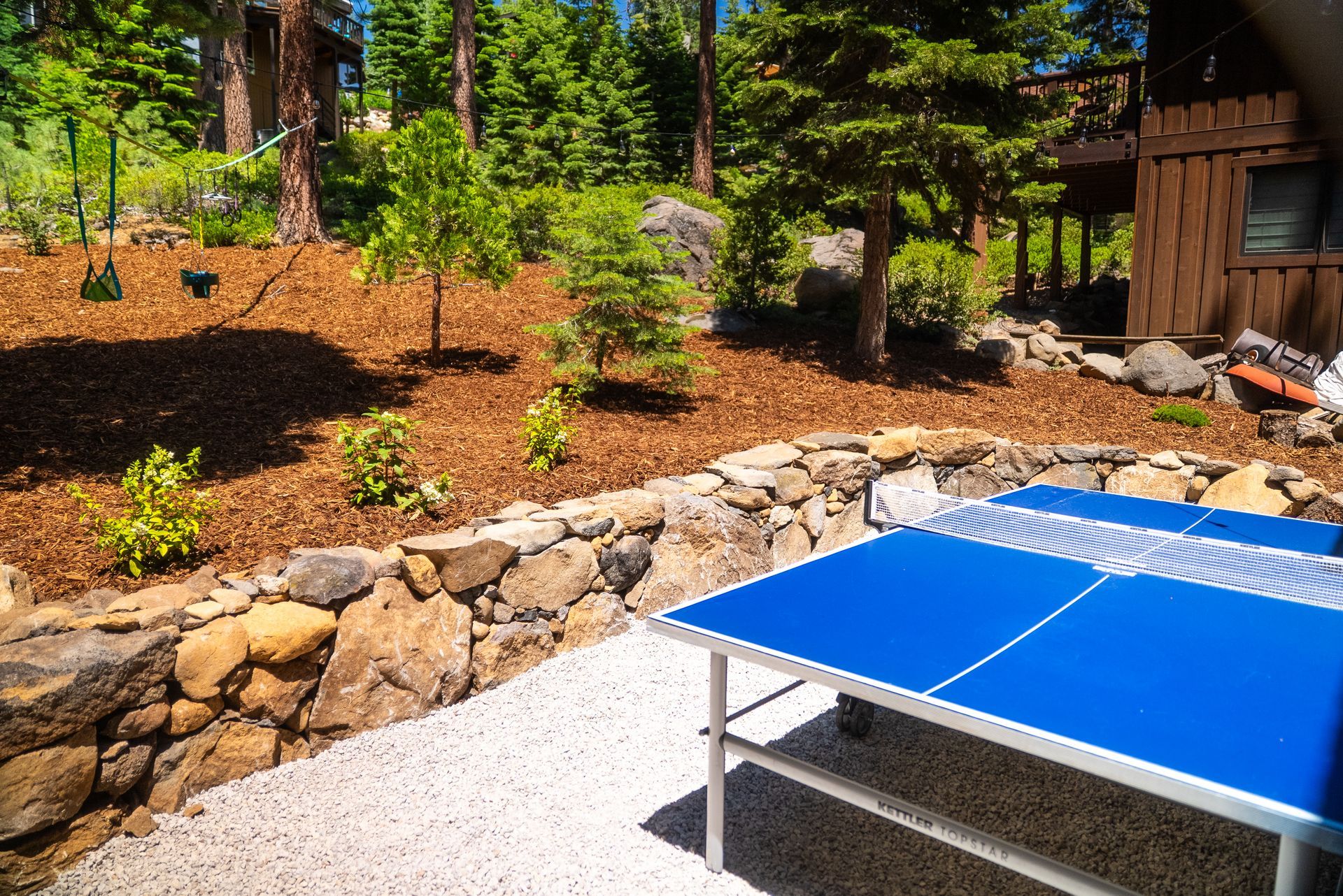 Outdoor ping pong table on a gravel patio with stone retaining wall. Trees and woods in the background.
