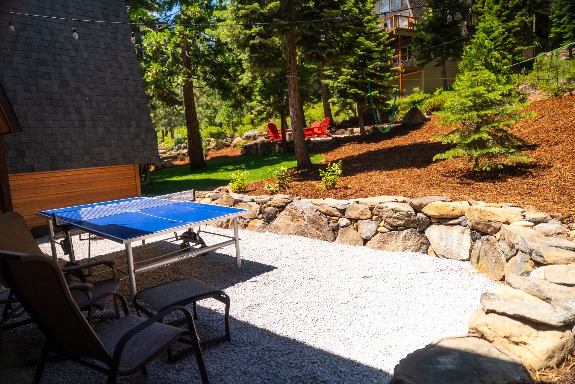 Patio with gravel surface, blue table, chairs, and stone wall, with trees in the background.