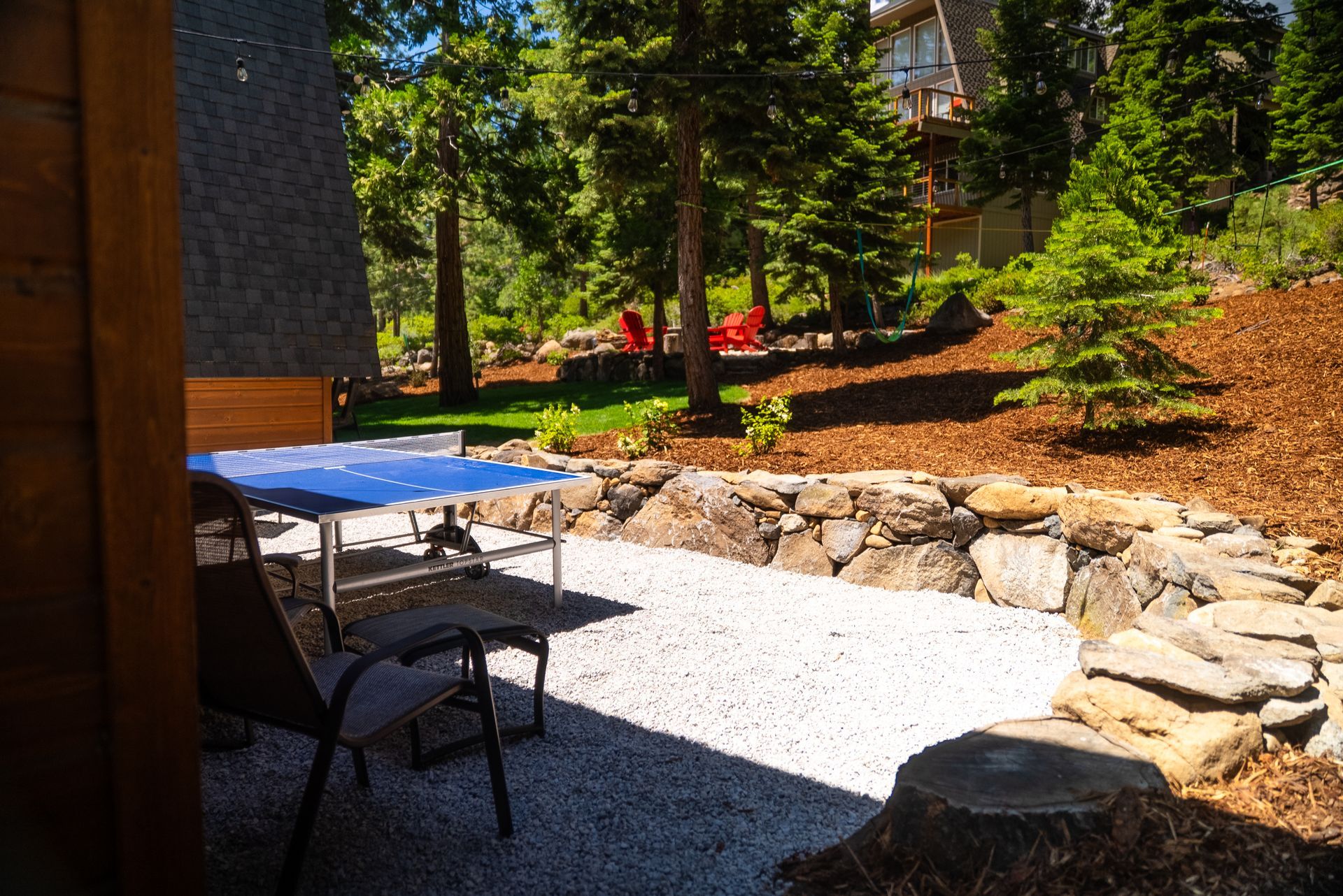Patio with gravel surface, stone wall, table, chairs, and landscaped hillside with trees.