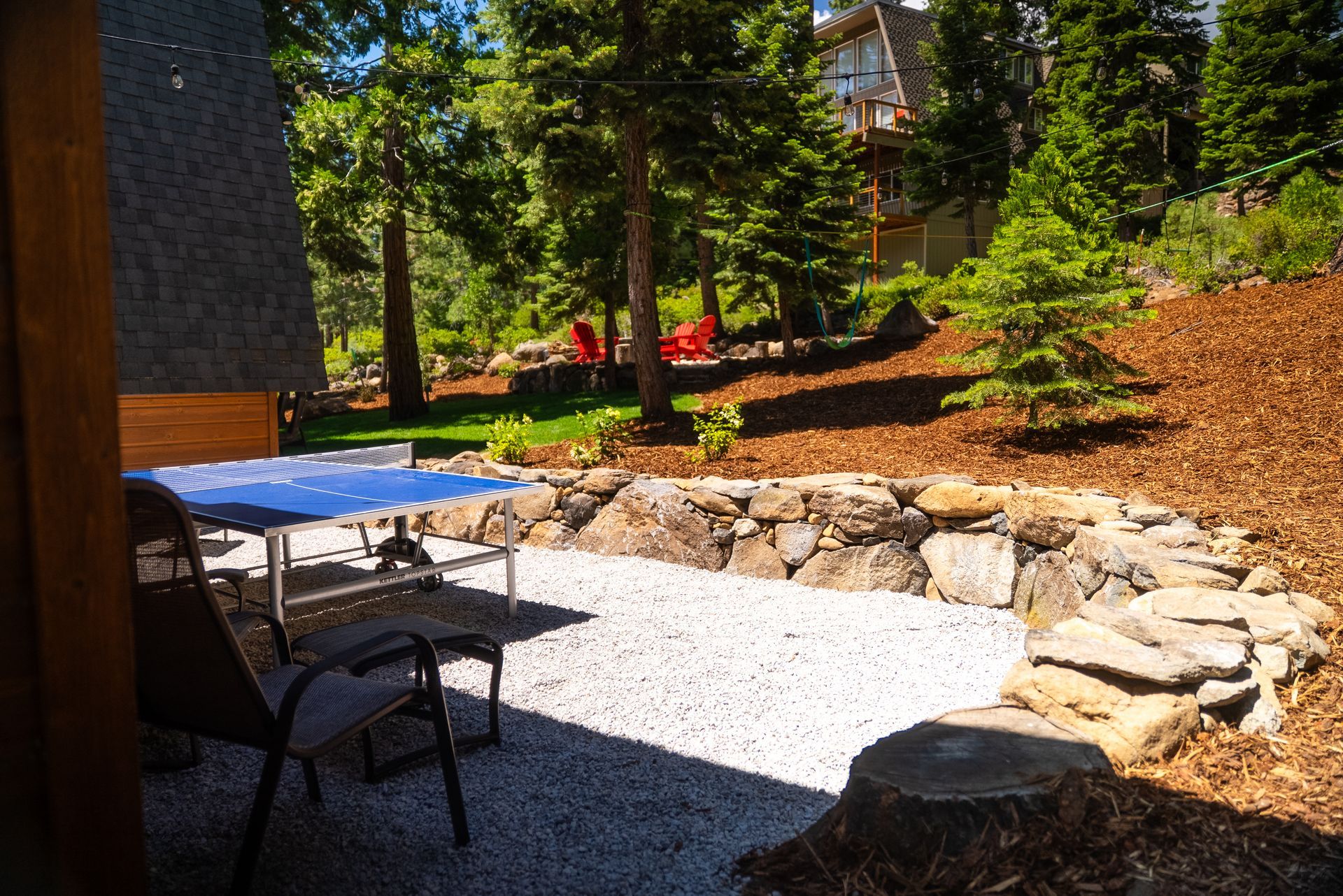 Patio with gravel, rock wall, table, chairs, trees, and red chairs in a sunny yard.
