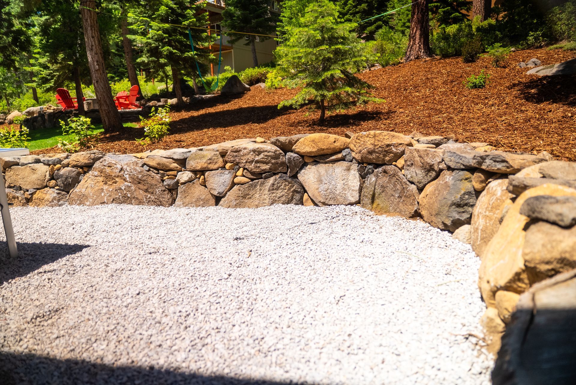 Stone retaining wall surrounds a gravel patio, with a backdrop of trees and wood mulch.