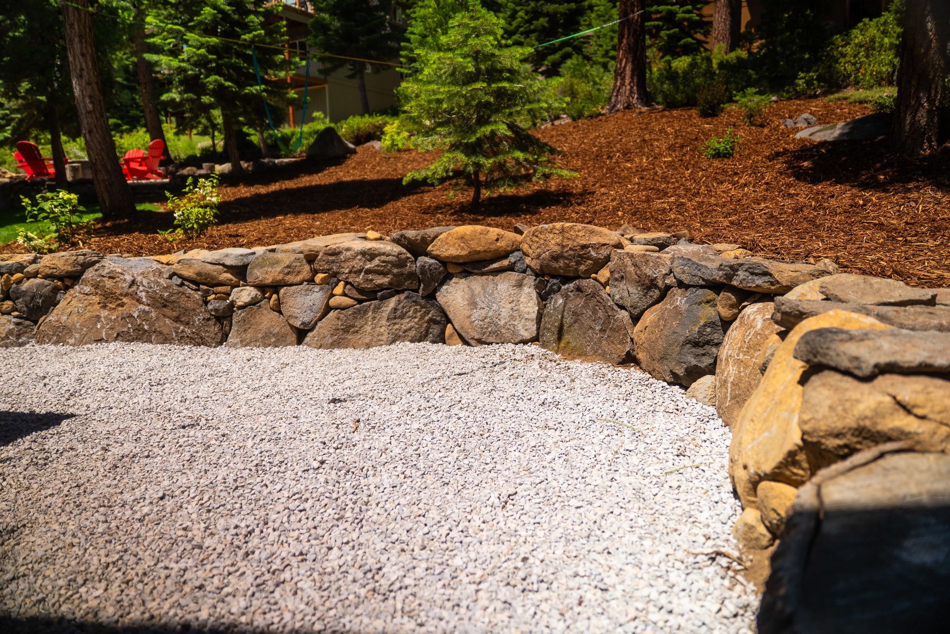 Stone wall borders a gravel area, with trees and mulch in the background.