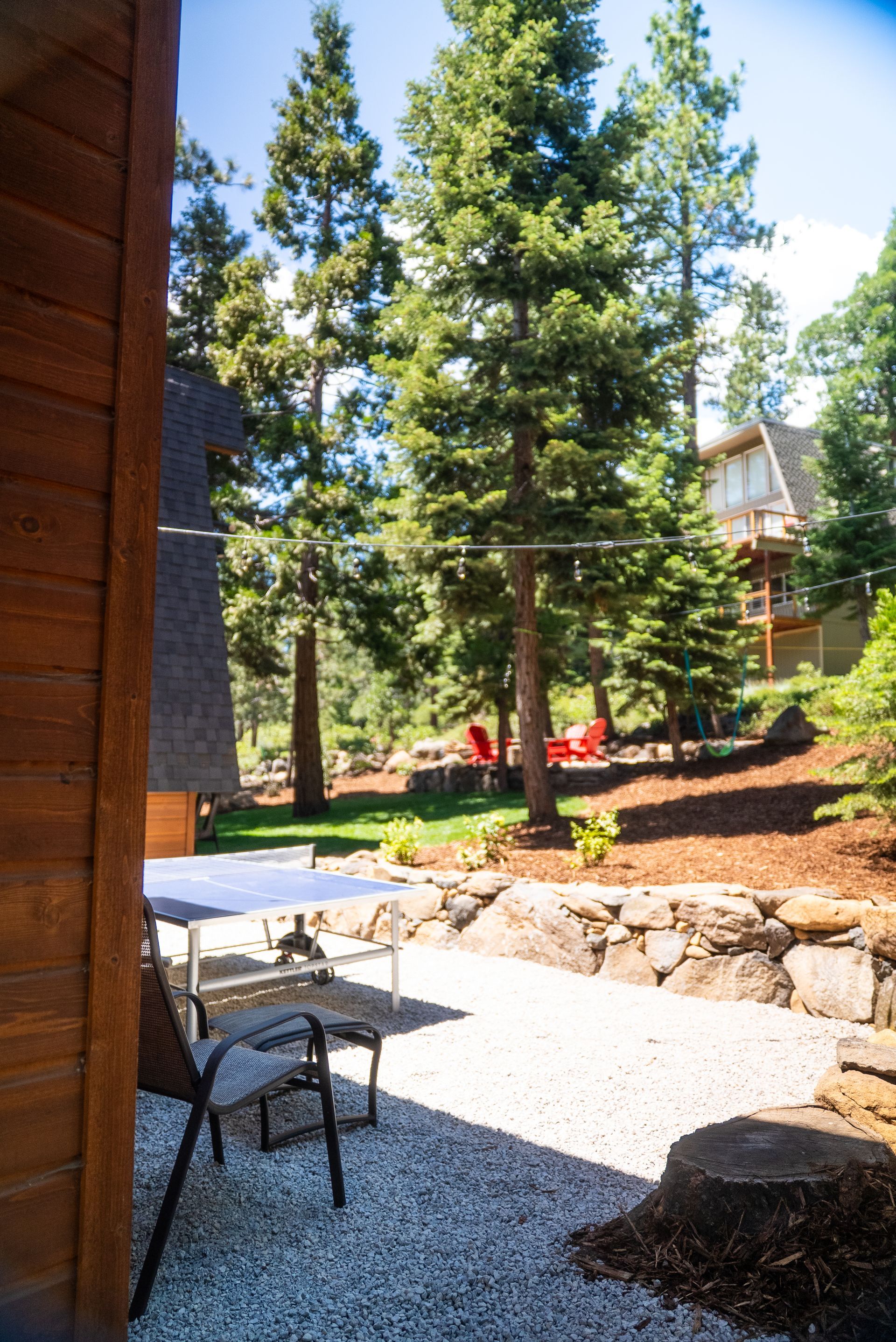 Patio with table and chairs, trees and a cabin in background. Sunny day.