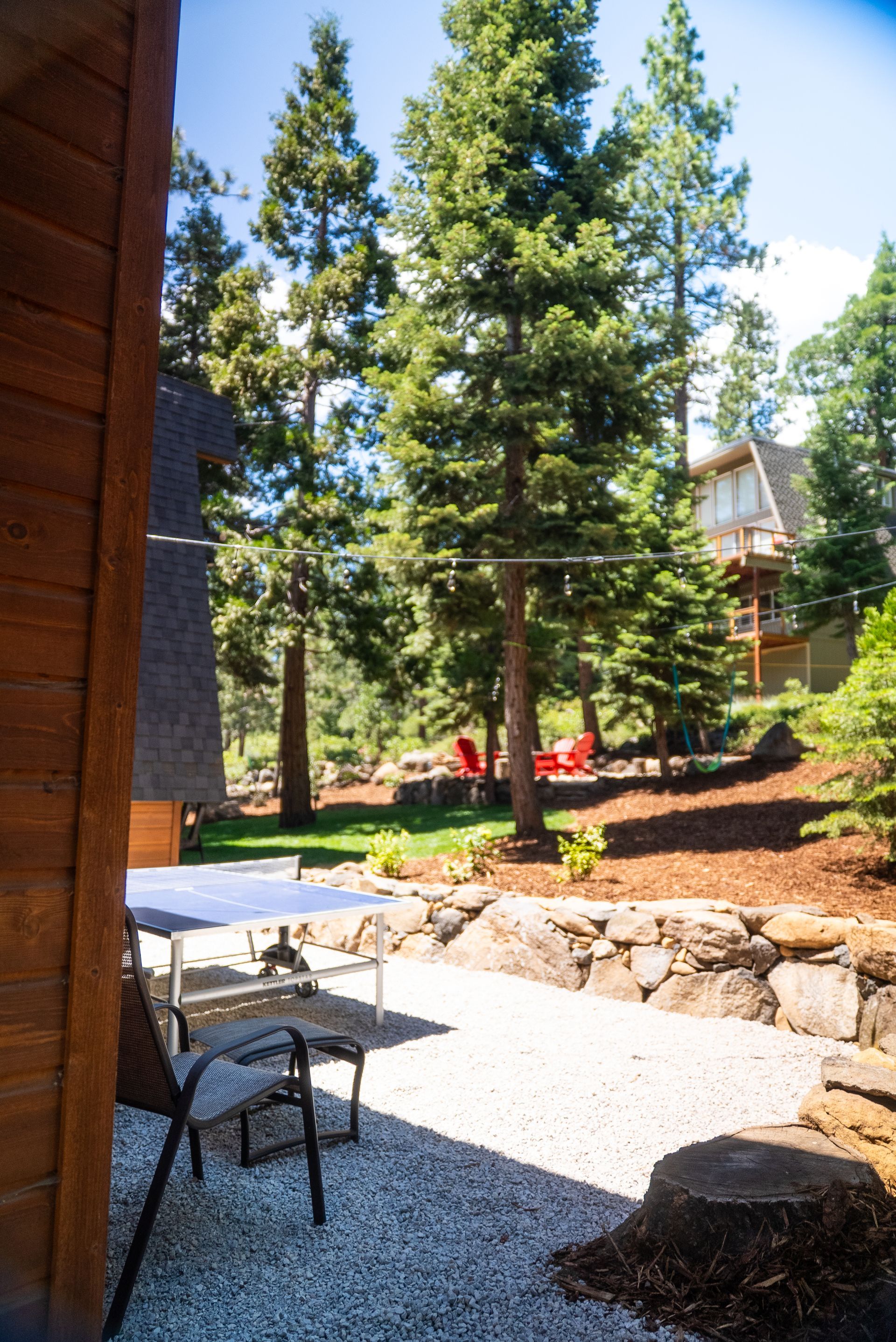 Patio with gravel, table and chairs. Backyard with trees and distant cabin on a sunny day.