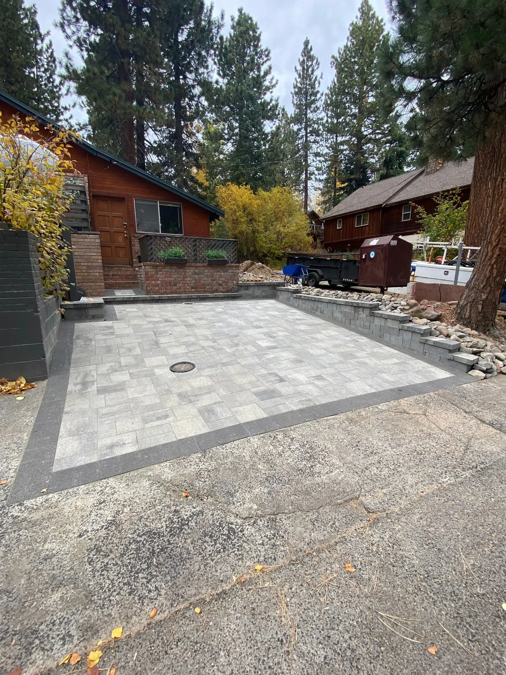 Gray paver driveway with dark border, leading to a wooden house with landscaping.
