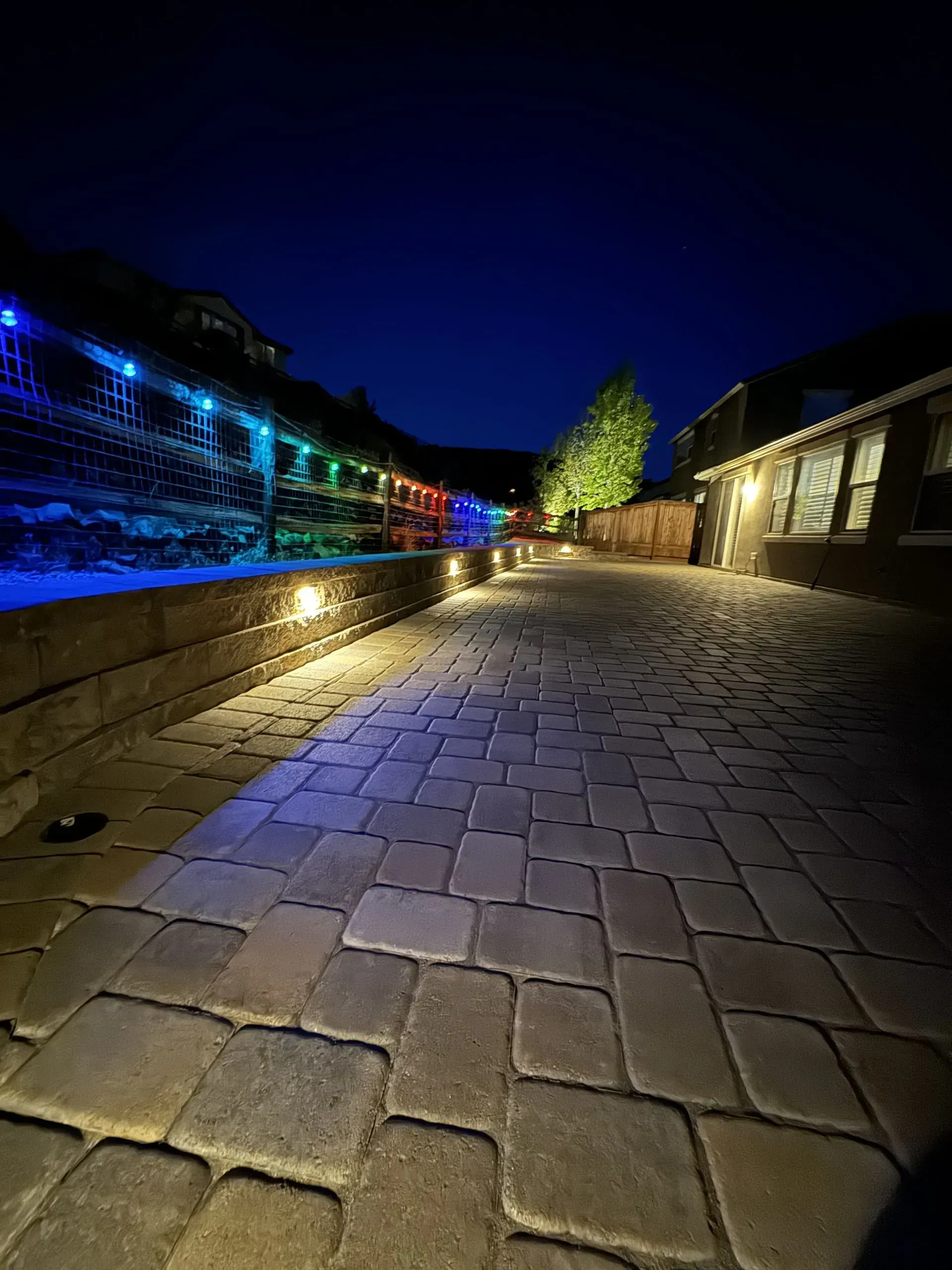 Paver pathway illuminated by lights at night, with colorful string lights on a wall in the distance.