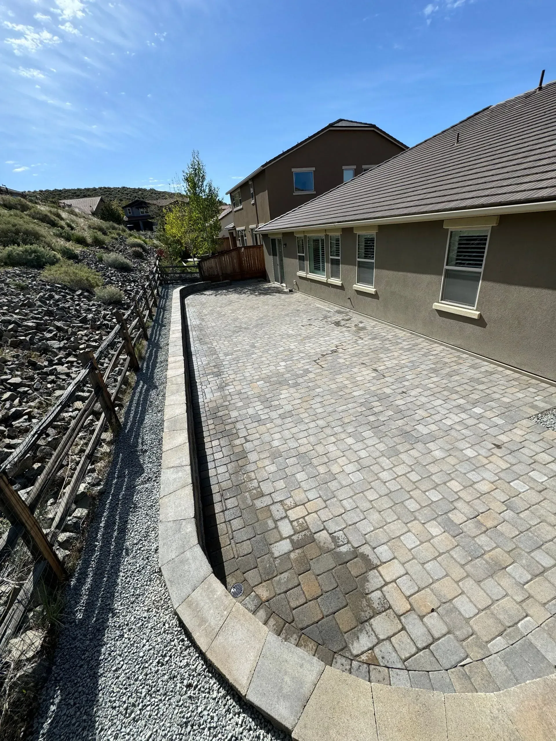 Paved patio next to a house with a gravel path and hillside in the background. Sunny day.