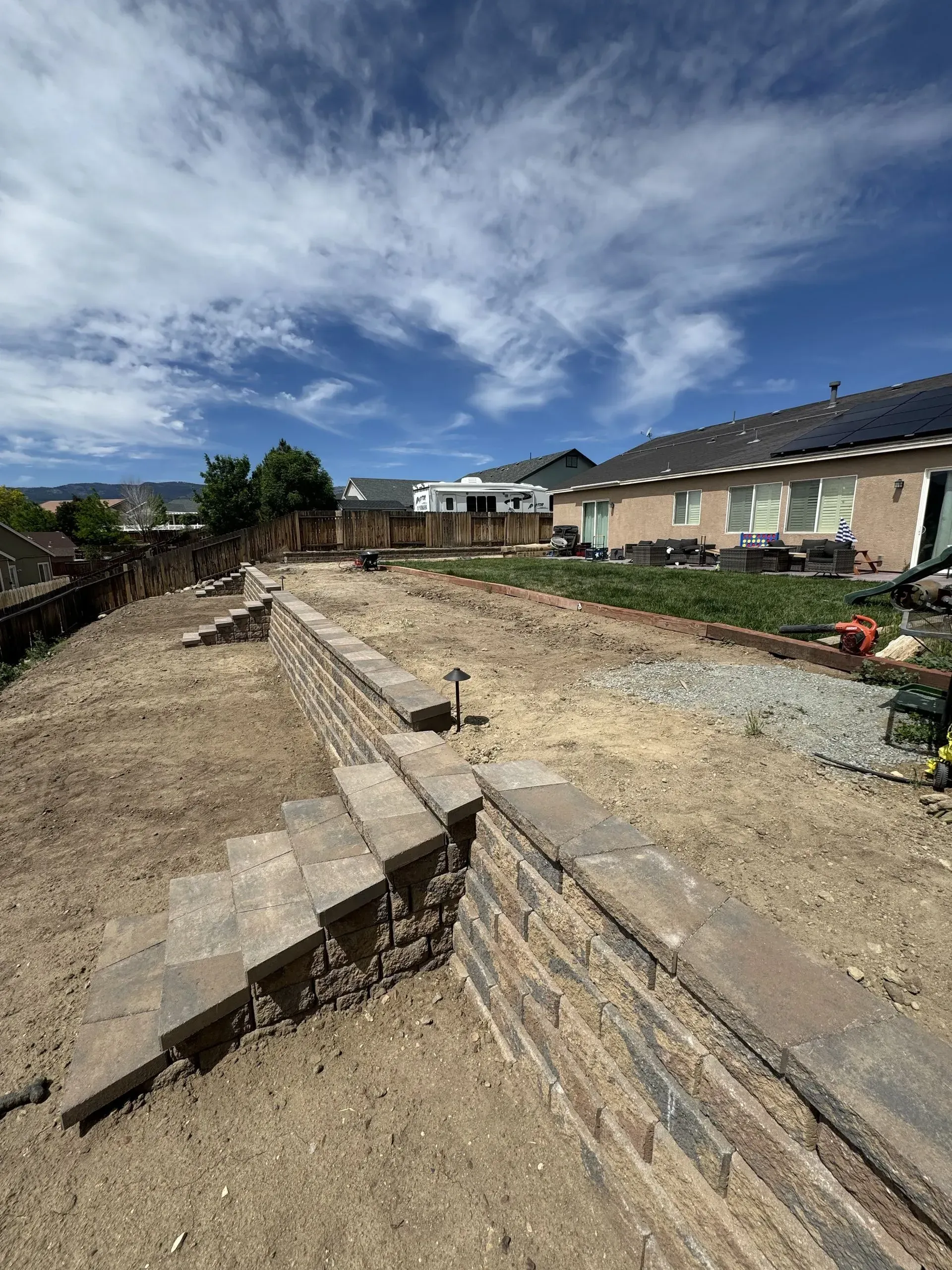 Backyard with tiered retaining walls, stairs, and exposed soil under a partly cloudy sky.
