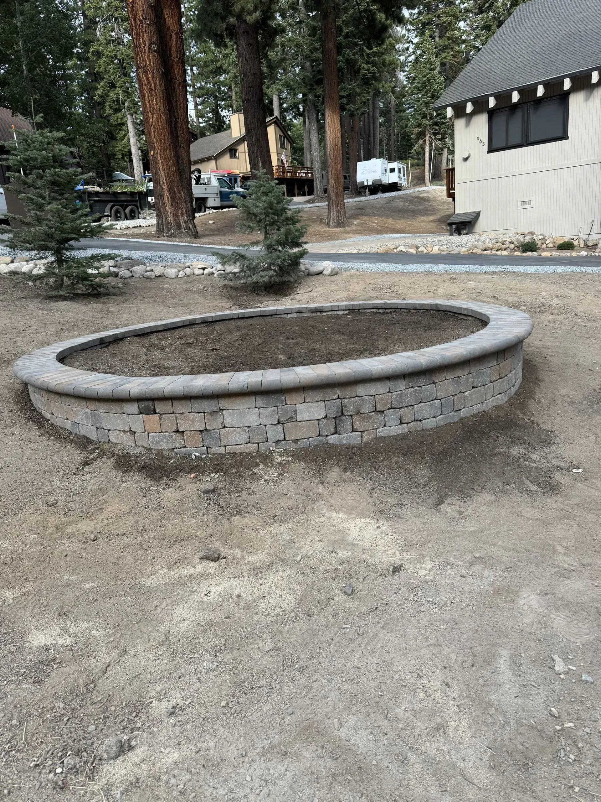 Oval-shaped brick planter in dirt; a building and trees are visible in the background.