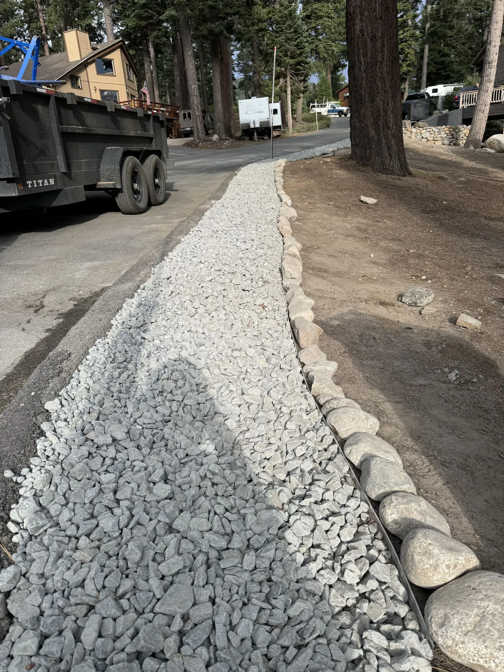 Gravel pathway edged with rocks, next to a paved road. Dump truck and trees are in the background.