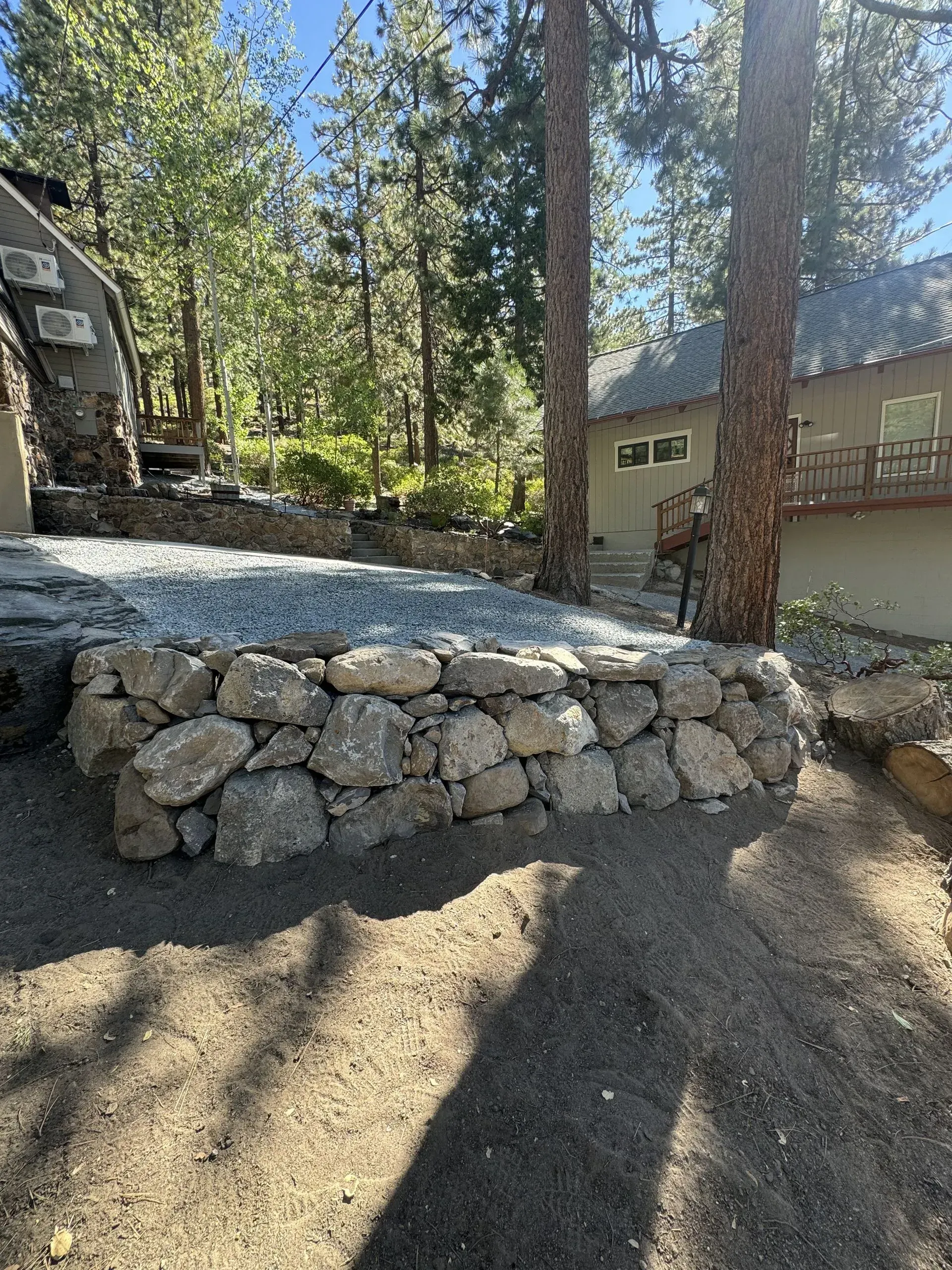 Stone retaining wall in front of a concrete patio, next to a beige house, with trees in the background.