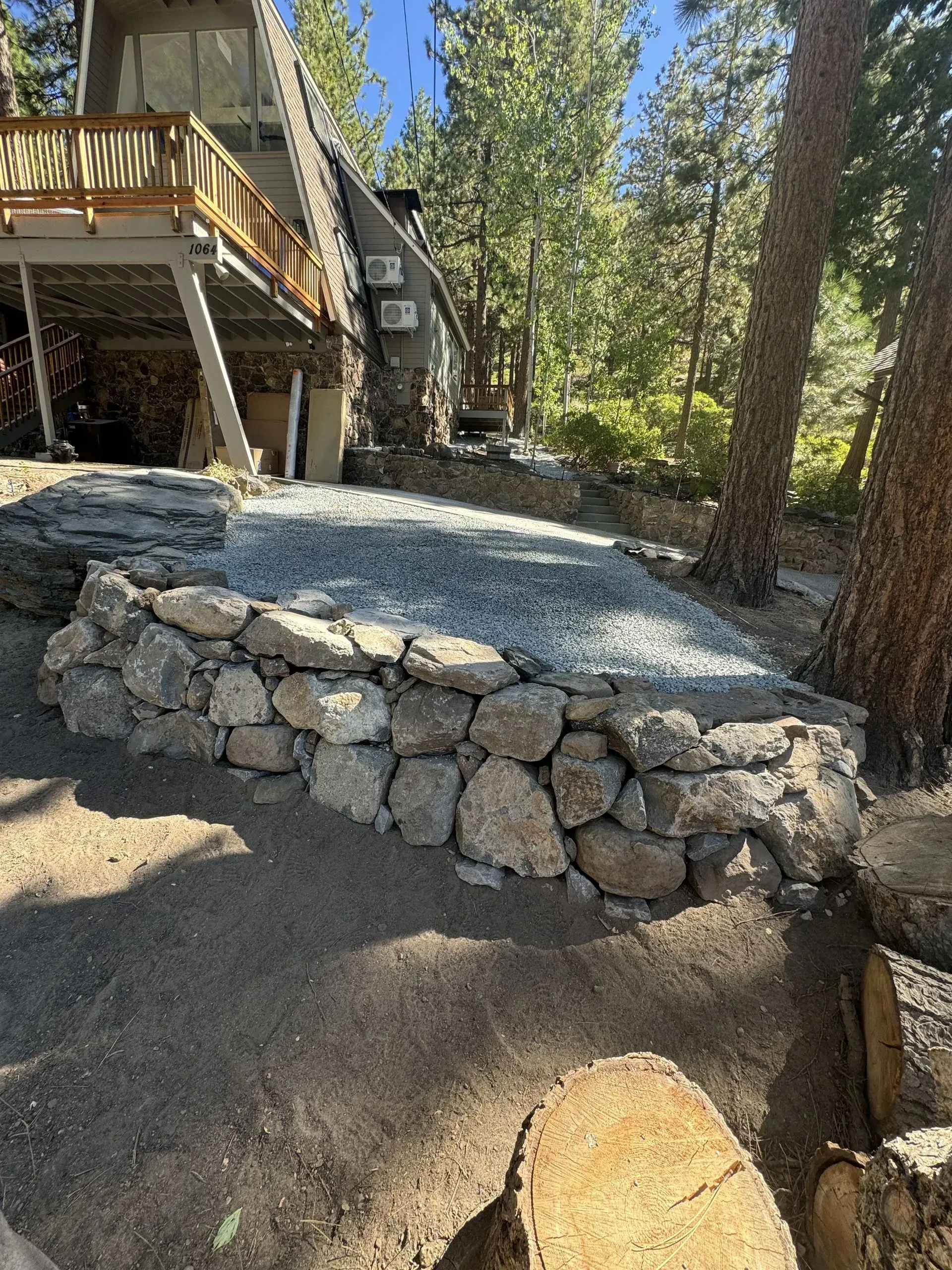 Stone retaining wall surrounds a gravel patio next to a house in a wooded area.