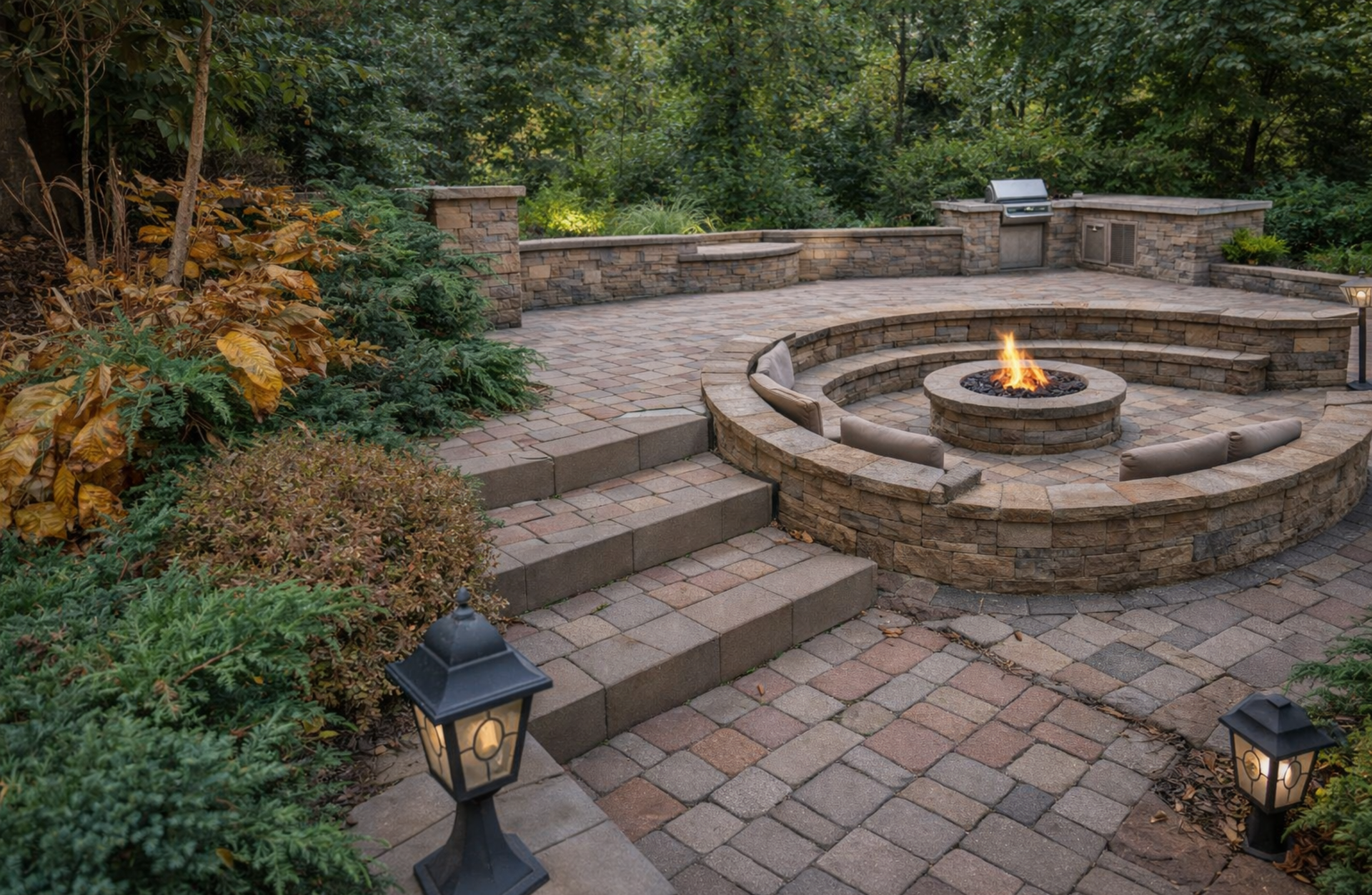A circular sunken fire pit area with stone seating and stairs in a paved backyard patio at dusk with outdoor lamps.