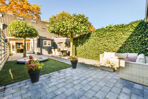 Backyard patio with grey pavers, green lawn, trees, hedges, and a wicker sofa.