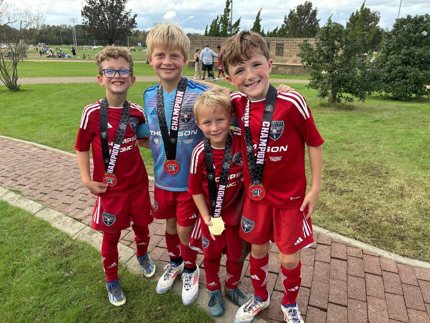Four kids in red soccer uniforms and medals standing on grass, smiling at the camera.