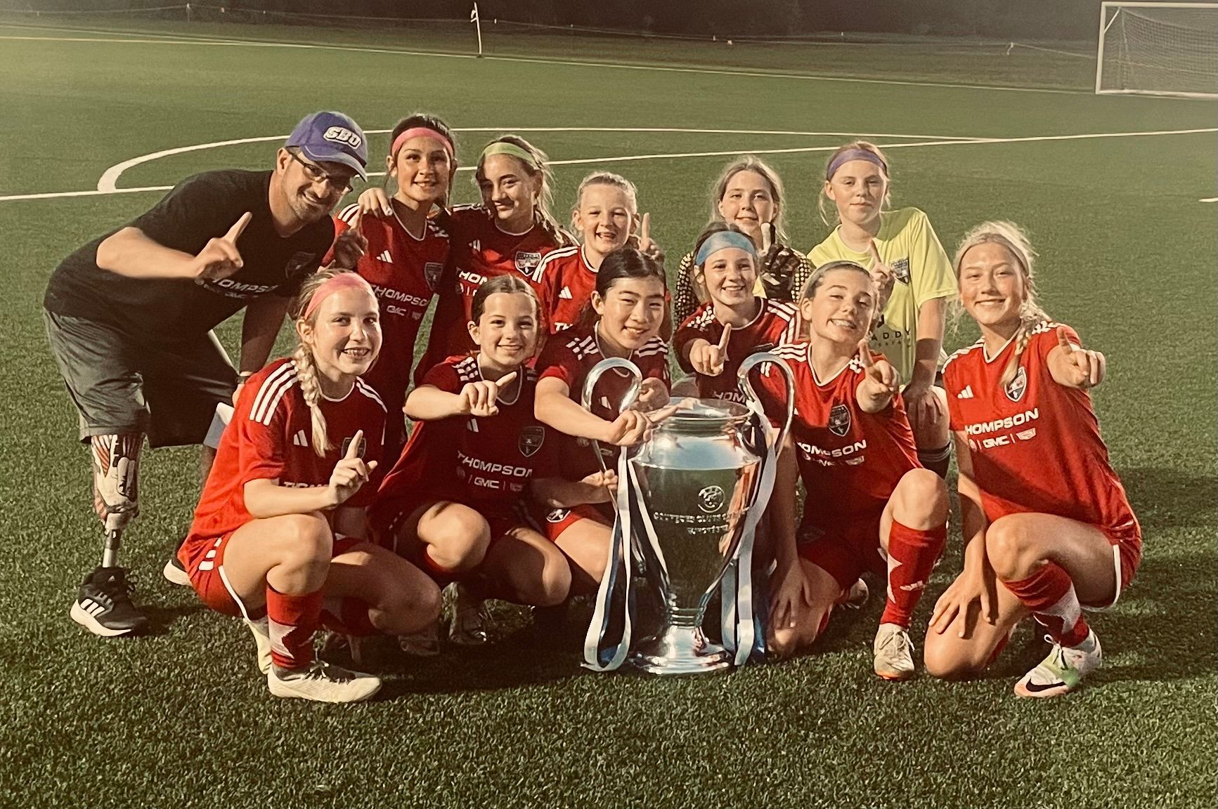 Soccer team posing with a trophy on a field at night, wearing red uniforms and smiling