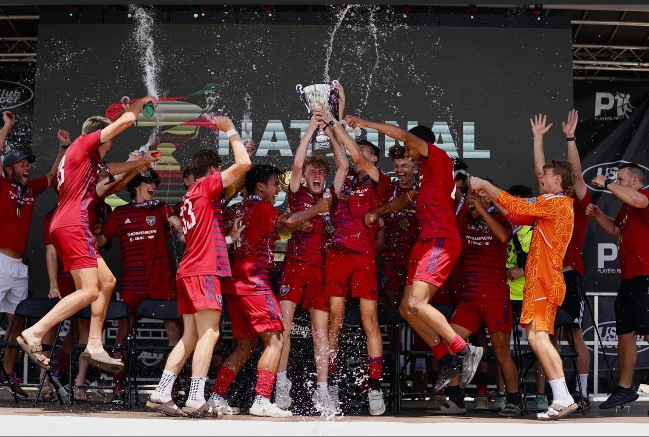 Soccer team in red jerseys cheering and lifting a trophy on stage with confetti falling