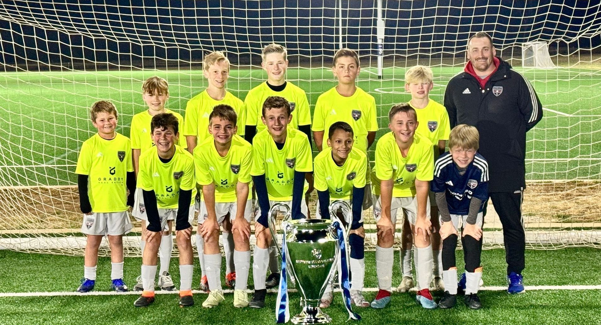 Youth soccer team posing on the field with a coach, goal net, and trophies at night