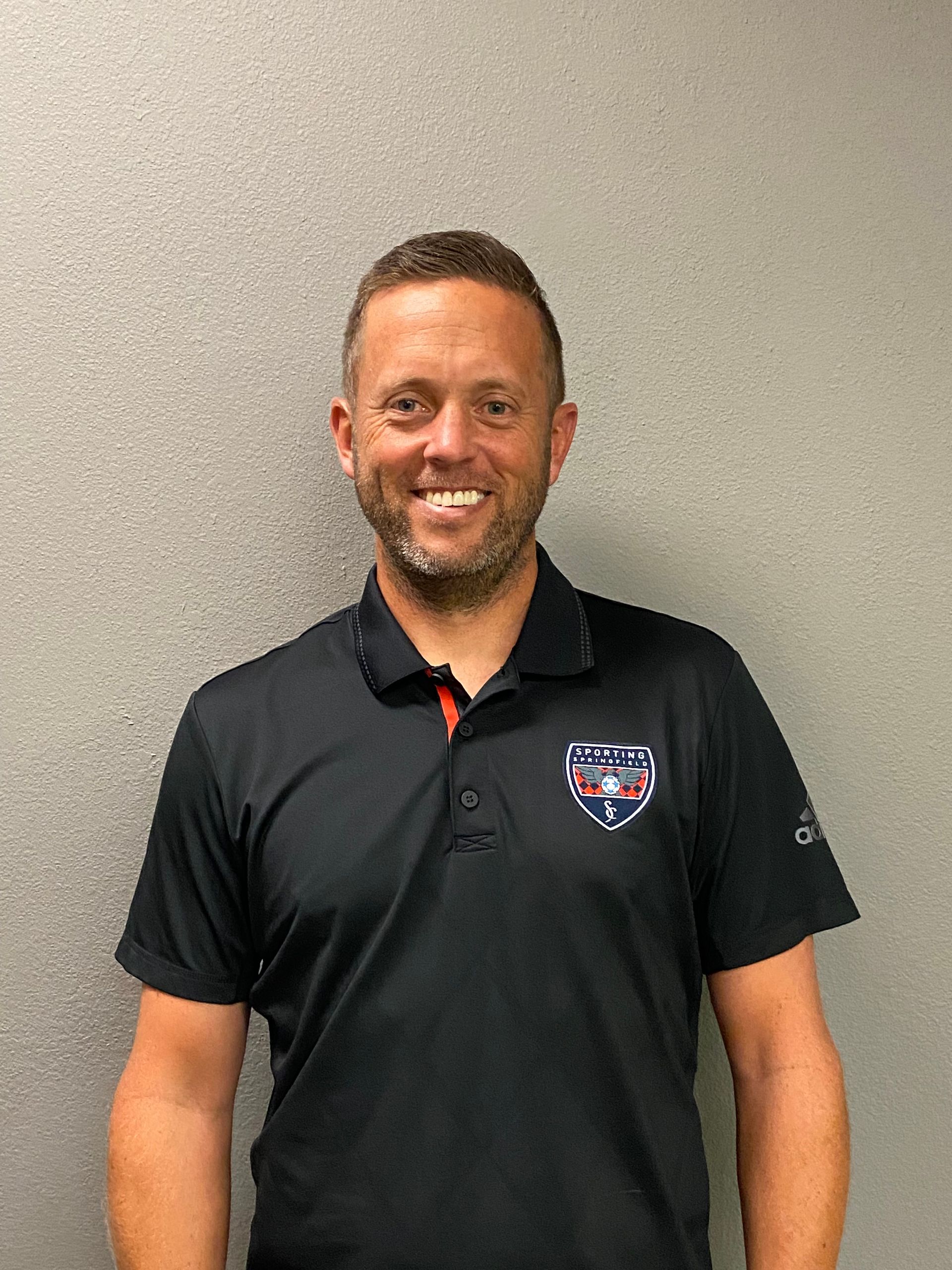 Smiling man in a black polo shirt standing against a plain beige wall.