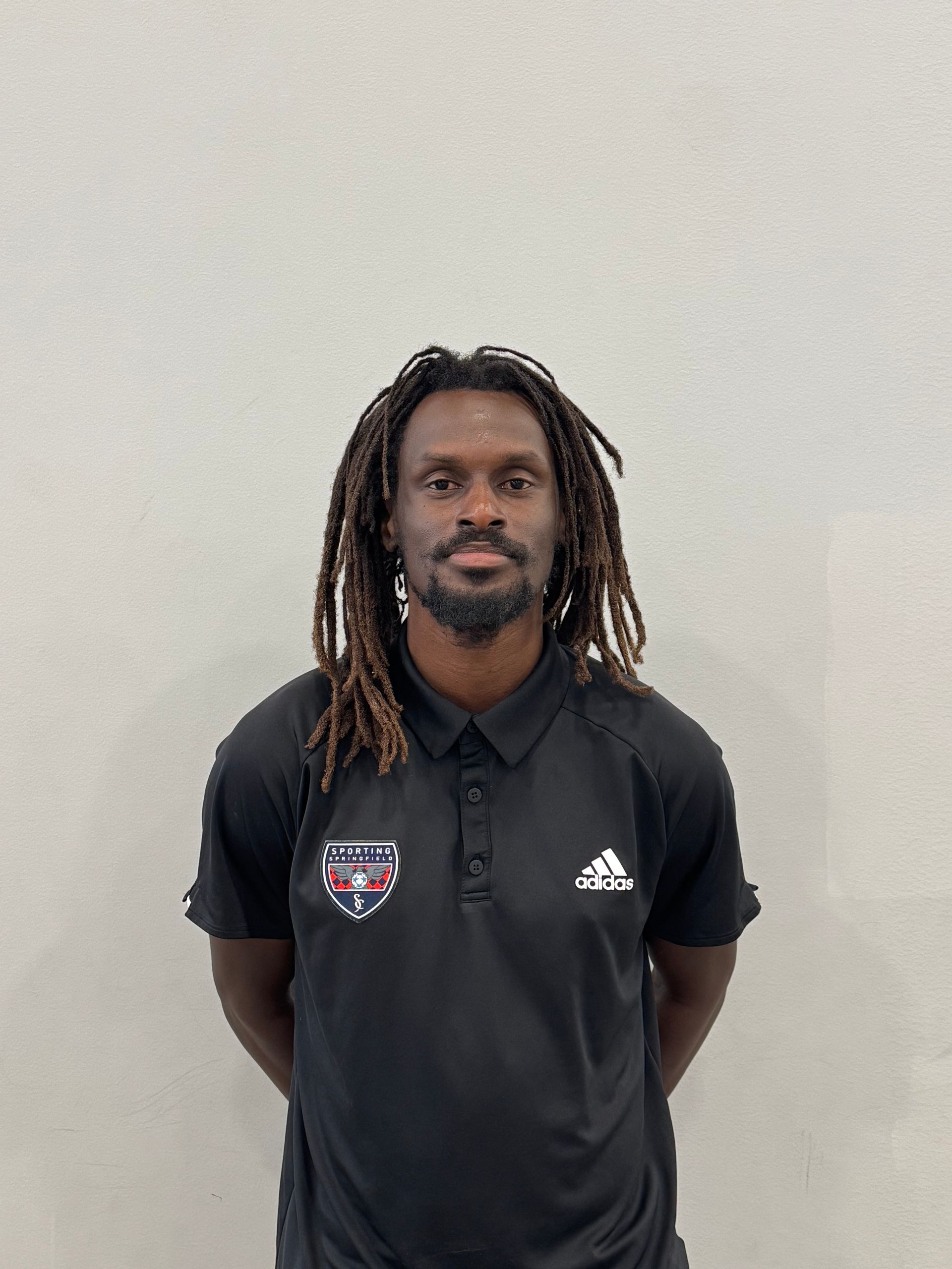 Man standing against a plain wall, wearing a black Adidas polo shirt with logo patches.