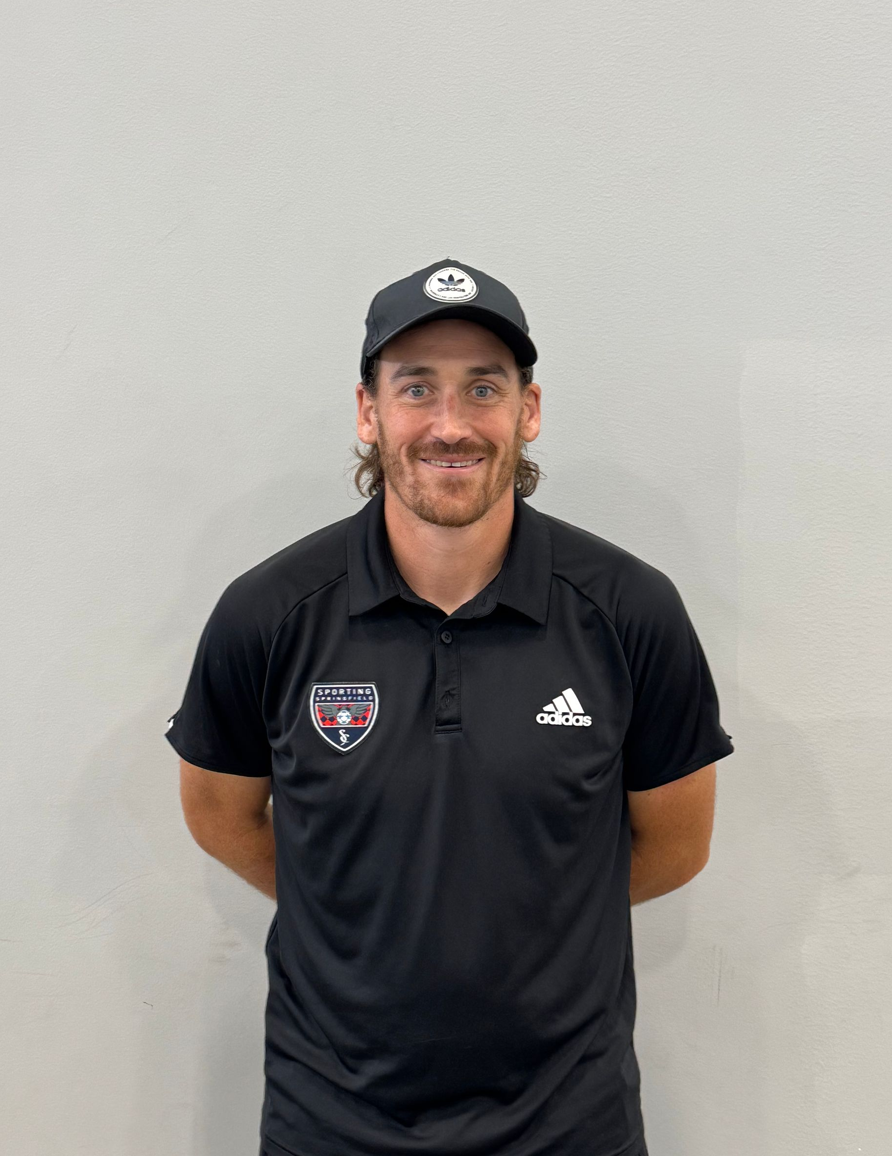 Man in black polo and cap standing against a plain wall, smiling at the camera.