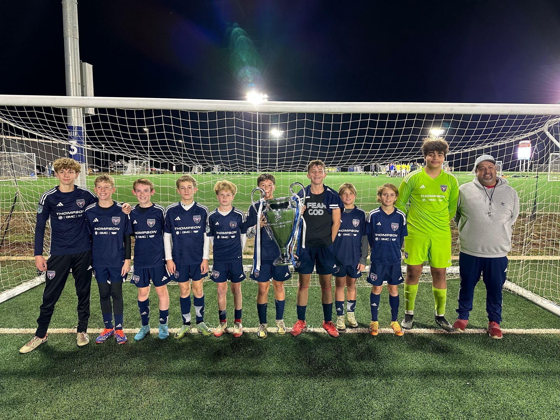 Youth soccer team lined up on a field at night in front of a goal, wearing navy uniforms and one neon green goalie kit