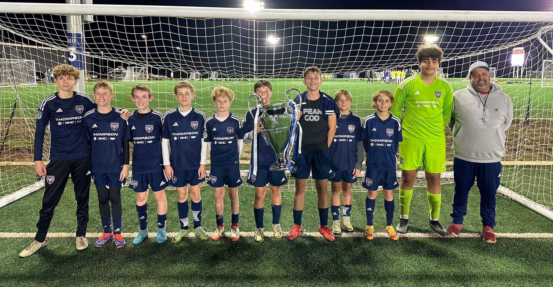Youth soccer team posing on a field at night, wearing navy uniforms with a neon green goalkeeper and coach.