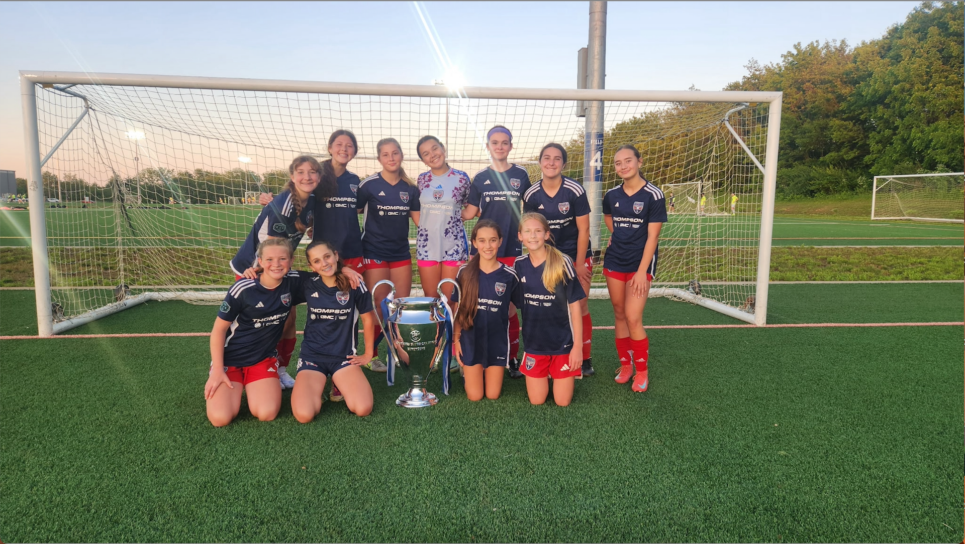 Youth soccer team posing on a field in front of a goal, holding a trophy and medals.