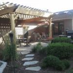 Backyard patio with wooden pergola, stone path, and lush shrubs beside a house