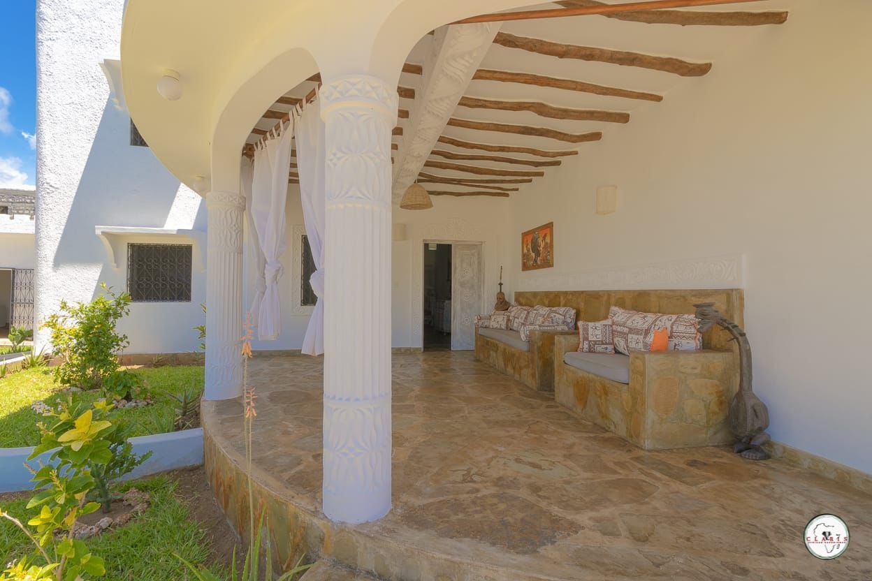 Covered patio with stone floor, white columns, wooden beam ceiling, and built-in seating in a bright outdoor area