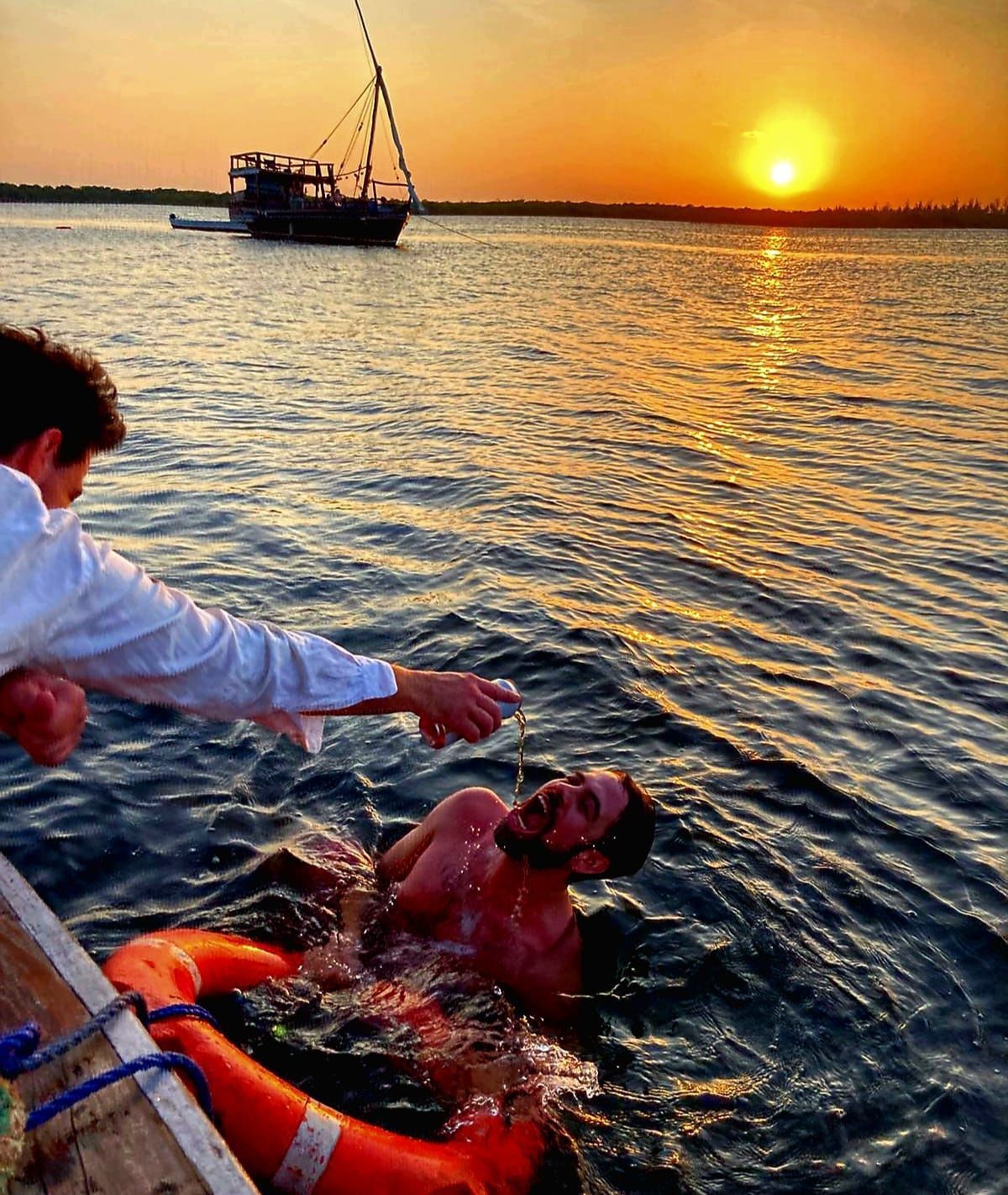 People rescuing a swimmer in the water at sunset, with a boat nearby.